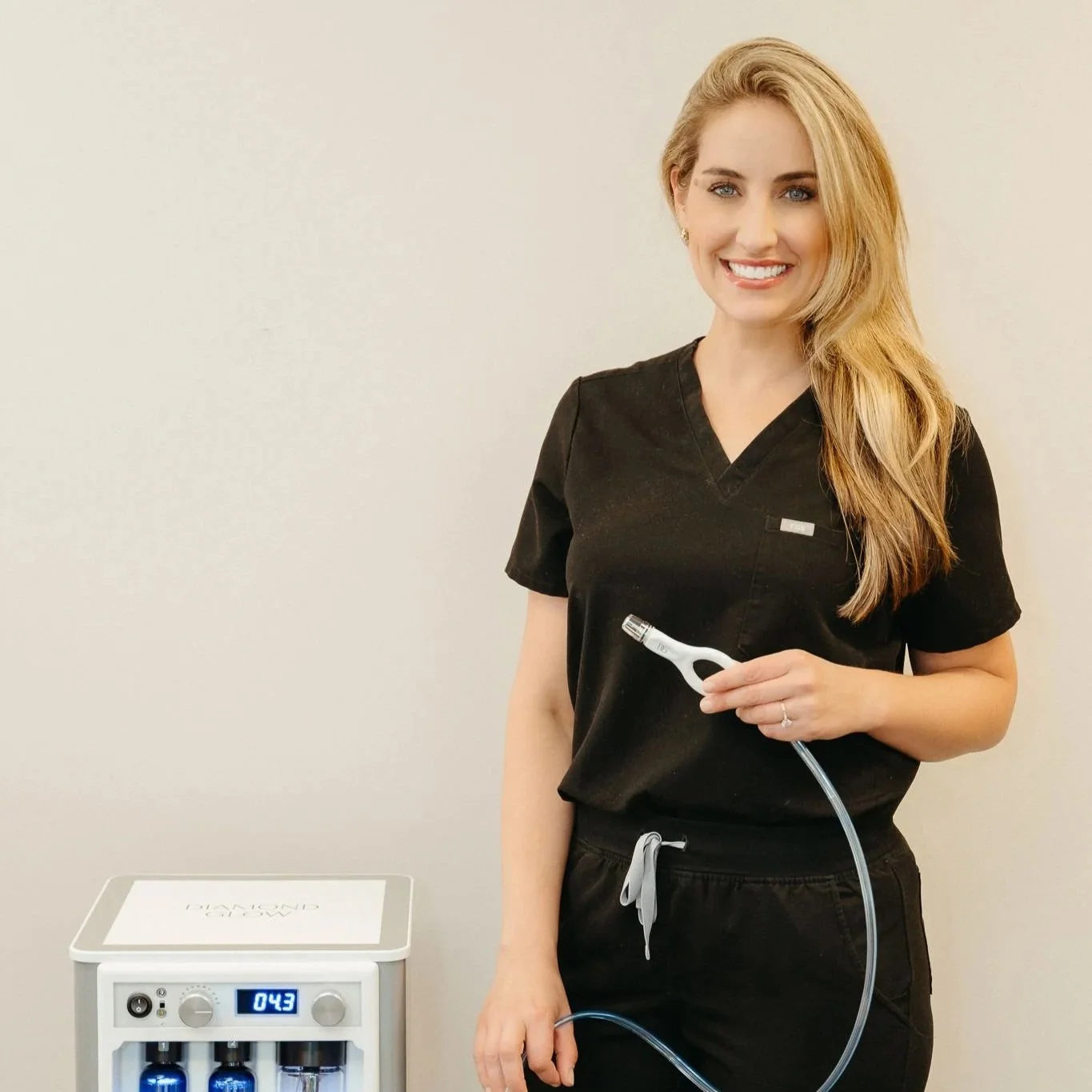 Aesthetician Christina - A woman in black medical scrubs holding a DiamondGlow device handpiece, standing next to a DiamondGlow device with a digital display reading 0.43 and three blue bottles inside.