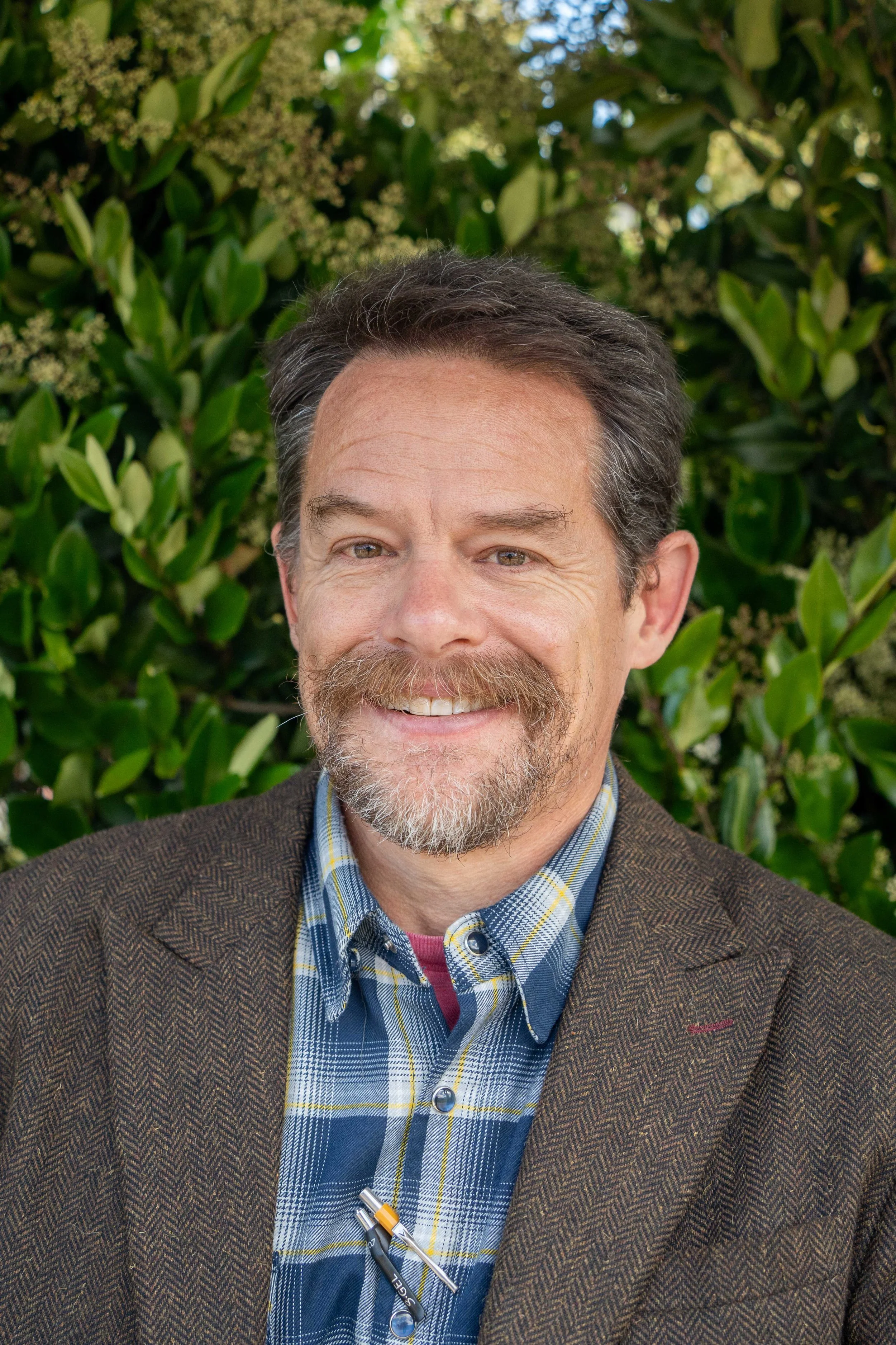 Portrait of a man wearing a gray blazer and white shirt, standing outdoors near a tree.