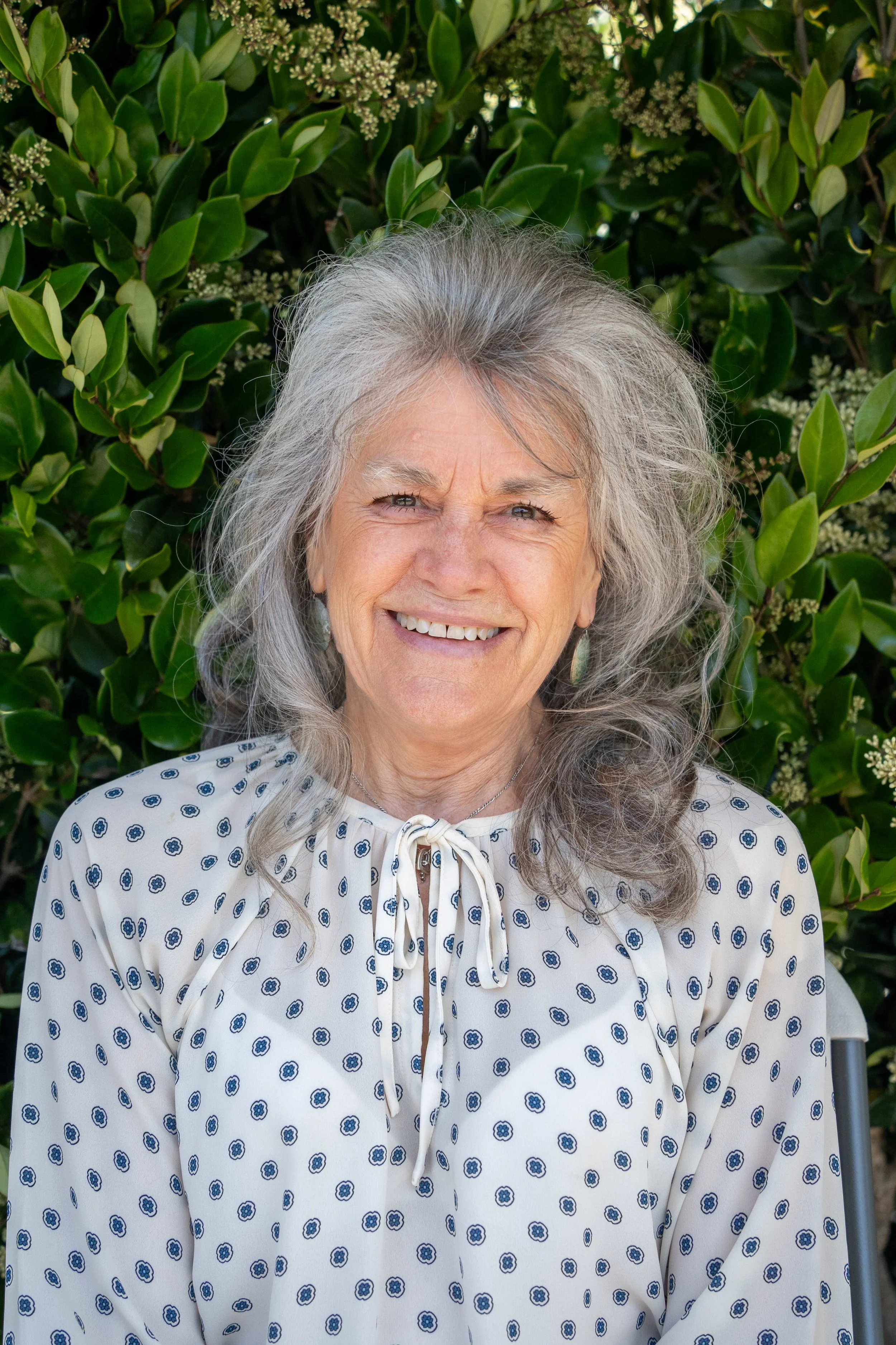 A smiling woman with long gray hair, wearing a navy blue polka dot shirt, stands outdoors against a large tree trunk and blurred green foliage in the background.