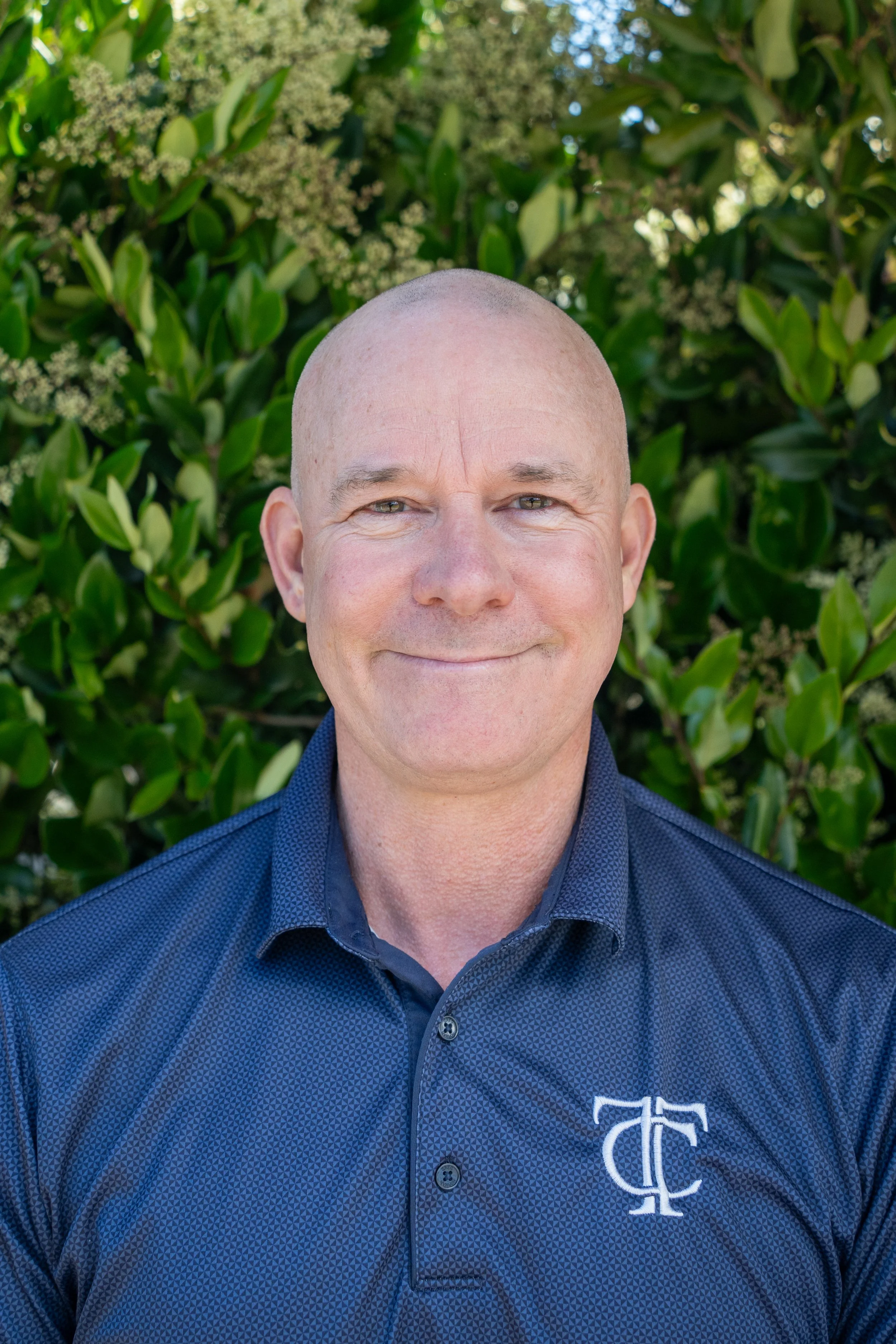 A bald man in a black polo shirt standing outdoors in front of a building with glass windows.