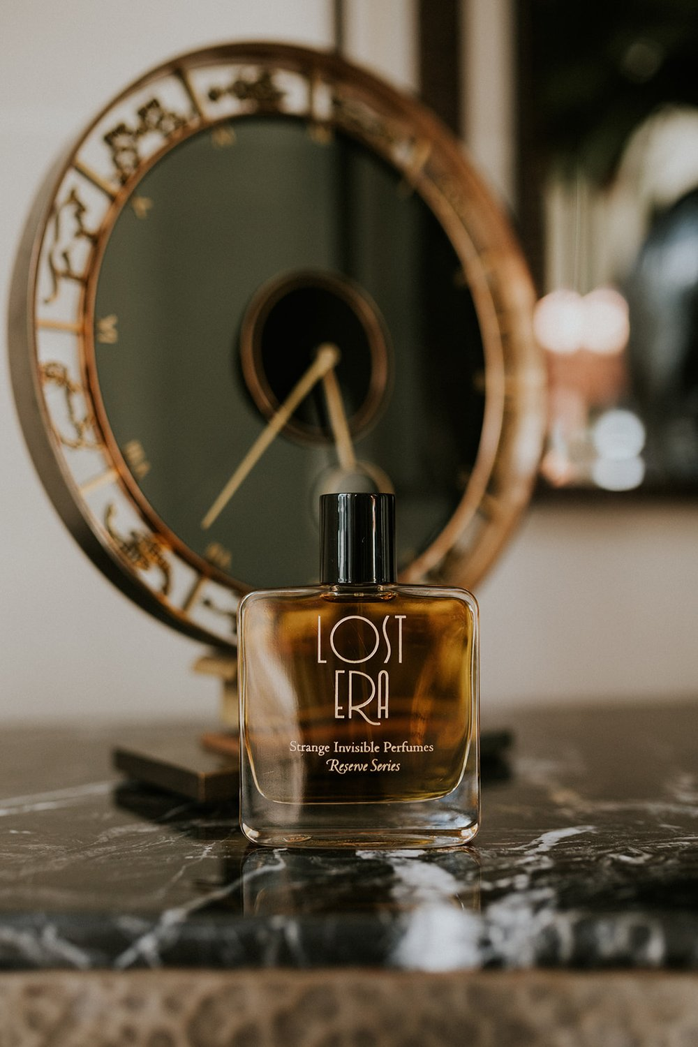 A bottle of Lost Era perfume placed on a marble table with a vintage clock in the background.