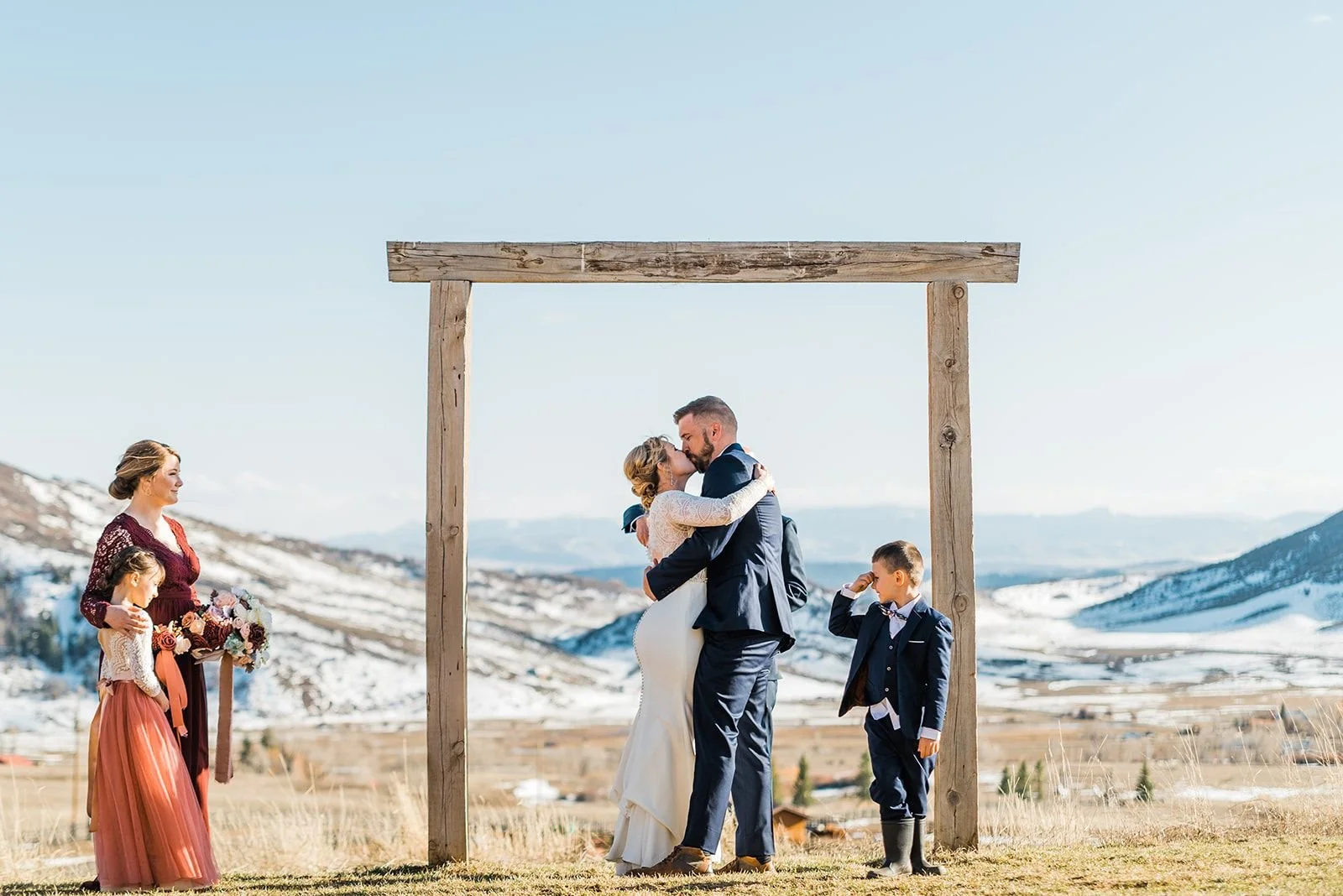 Bride and groom kissing with mountains in background and little boy hiding his eyes during their winter elopement at La Joya Dulce in Denver, Colorado.