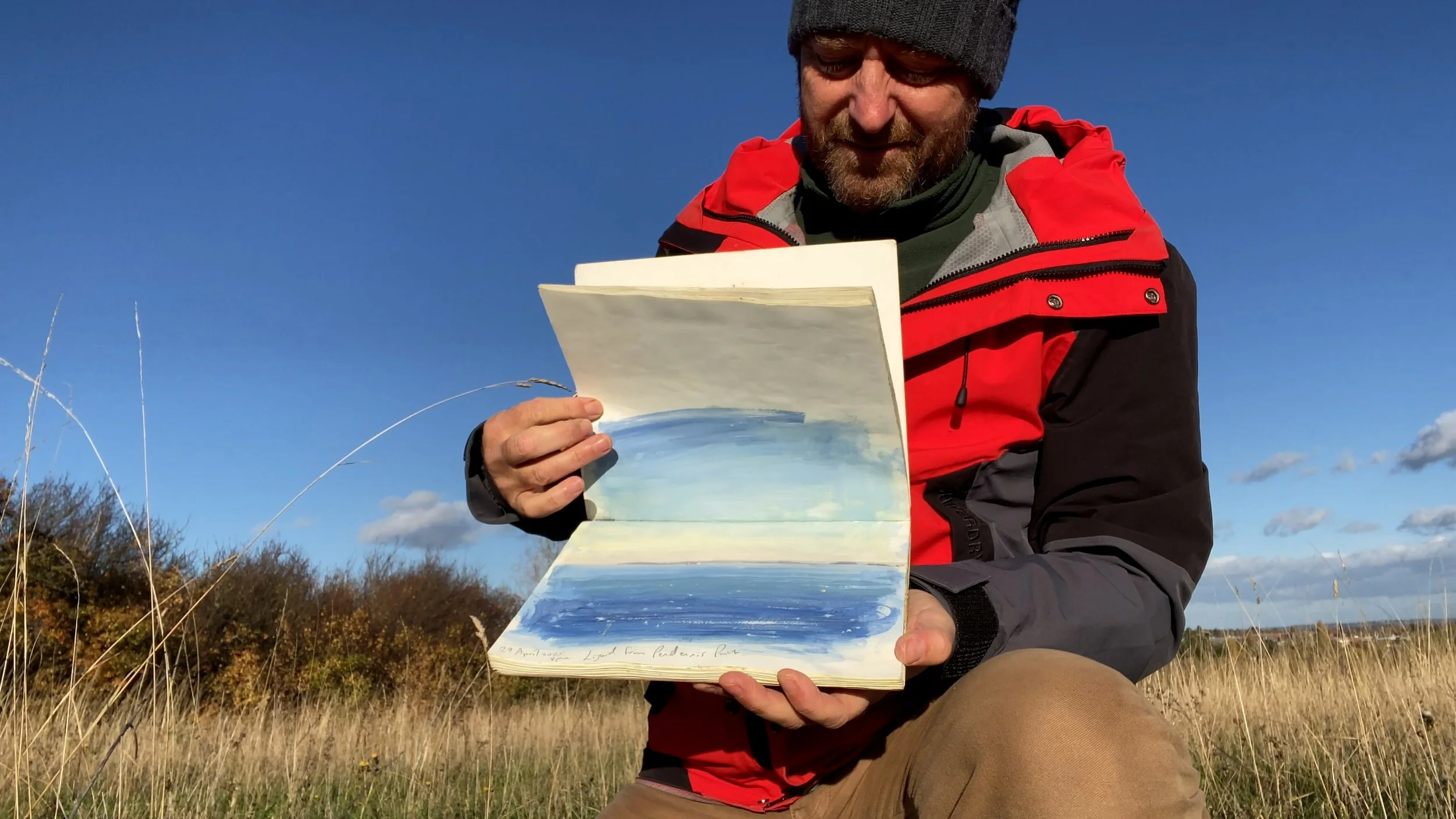 A man wearing a red and black jacket and a gray beanie is sitting outdoors in a grassy field, holding an open sketchbook with a watercolor painting of a landscape, and looking at it.
