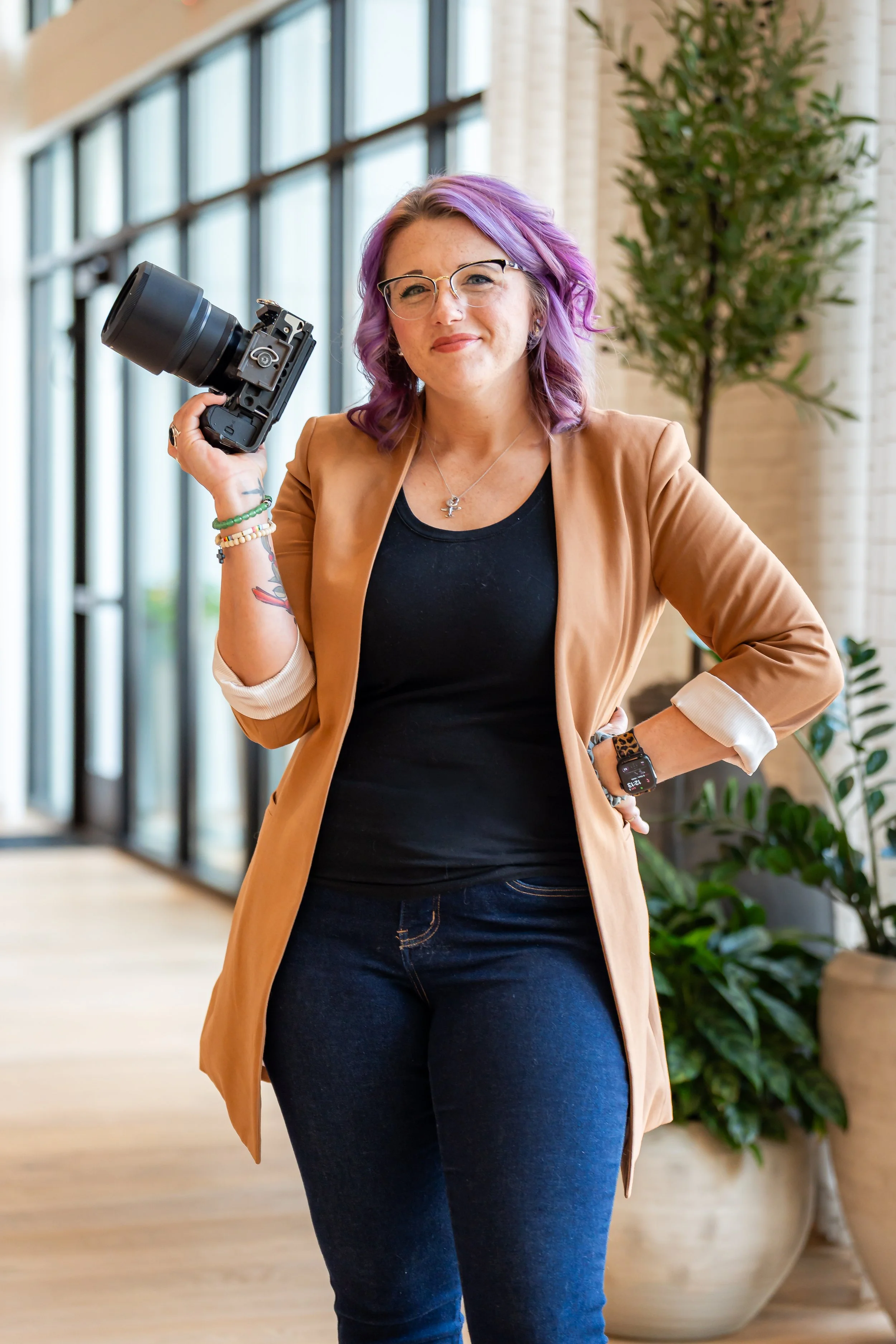 A woman with purple hair and glasses holding a camera, standing in a modern indoor space with large windows and potted plants,  image of Lauren Kress of Lauren Liz Photo.