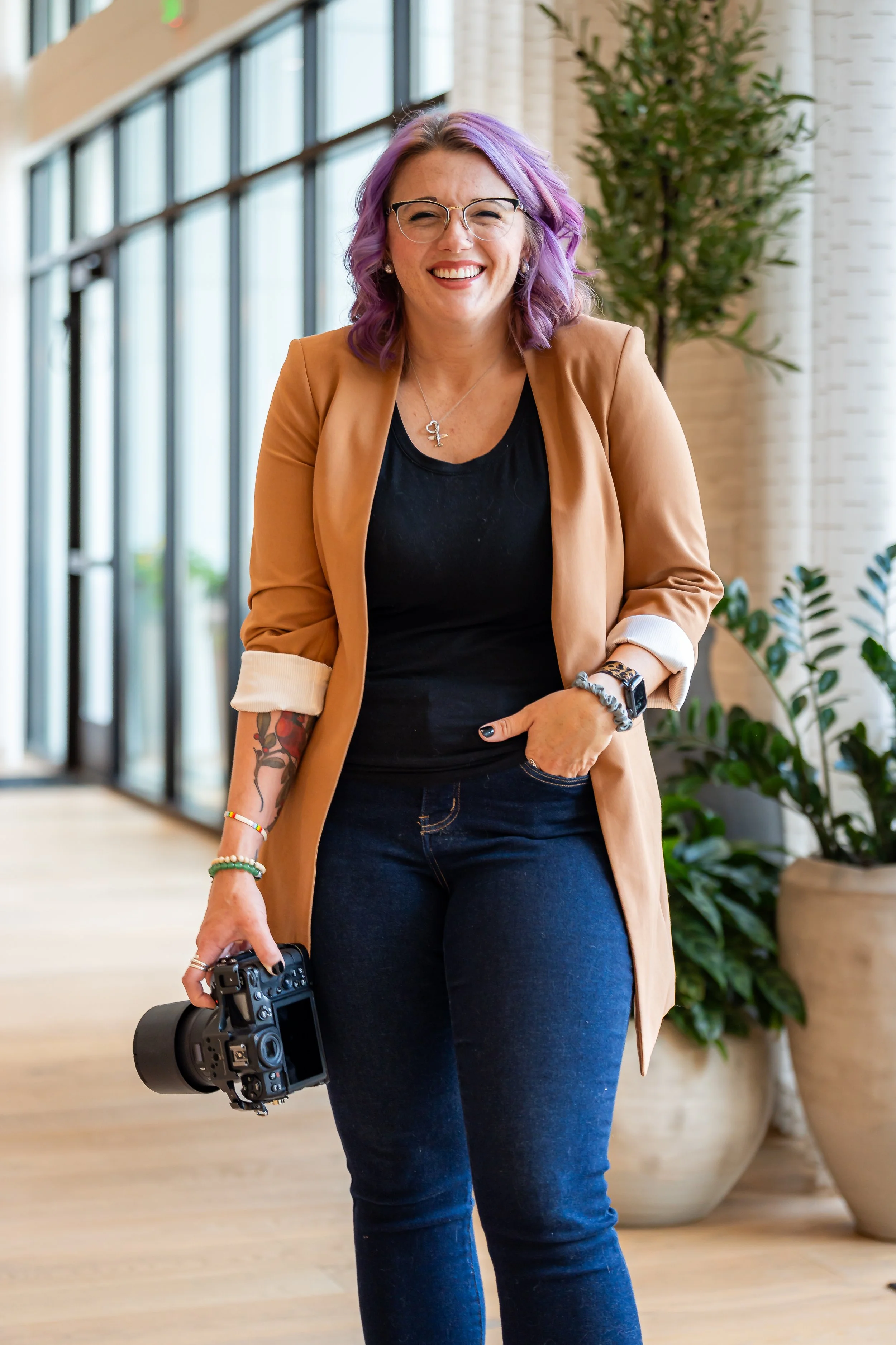 Lauren Kress, a woman with purple hair, glasses, and tattoos, smiling while holding a camera in a bright, modern indoor space with large windows and potted plants.