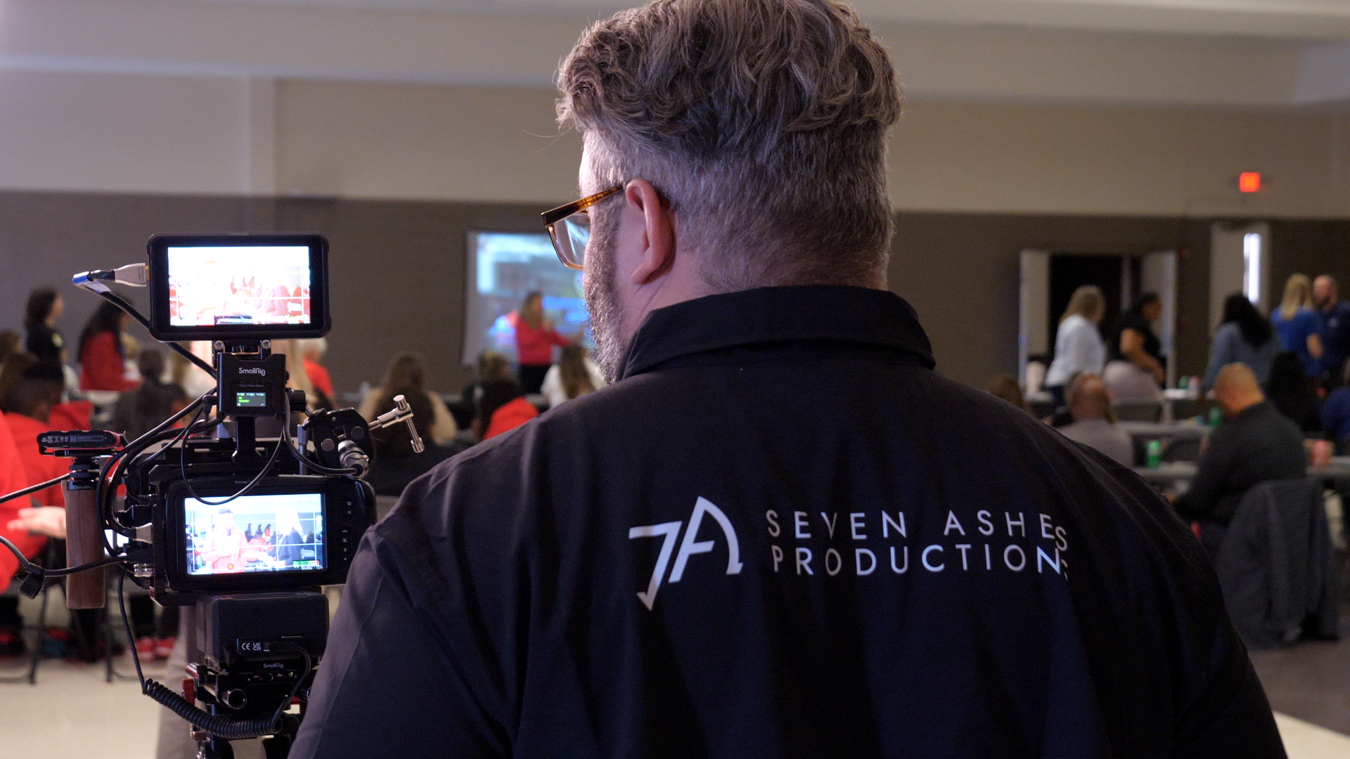 A man with short curly hair, glasses, and a beard operates a professional camera at an indoor event. The man wears a black jacket with the logo and name 'Seven Ashes Production' on the back. In the background, a group of people are seated and standing in a large room, possibly a conference or seminar.