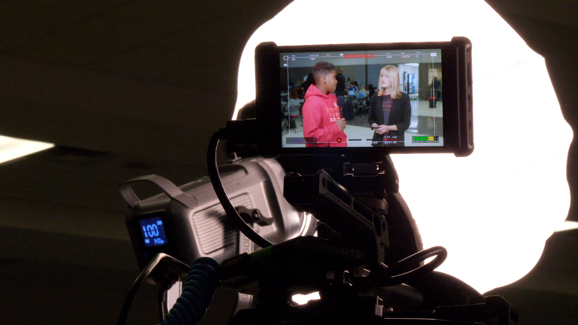 A camera recording an interview between two women in a public space, with one woman holding a microphone and the other woman holding a phone.