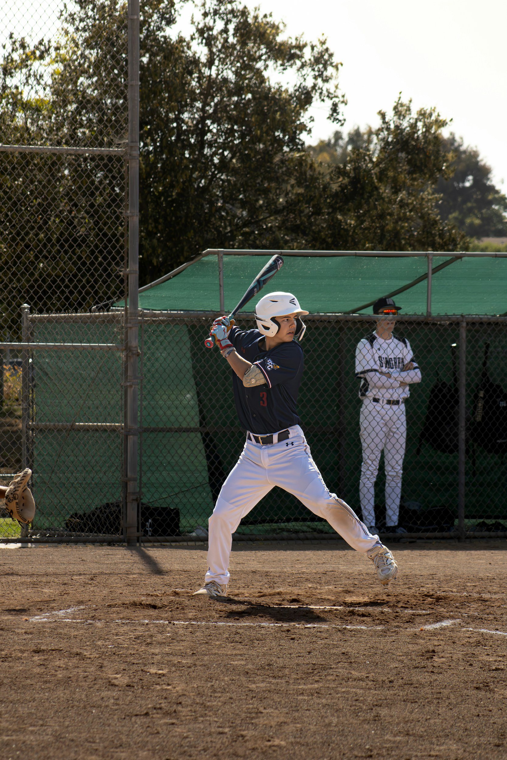 A baseball player in a dark uniform and white helmet at bat, standing in the batter's box on a dirt field. A teammate in a white uniform and cap stands behind a chain-link fence in the background.