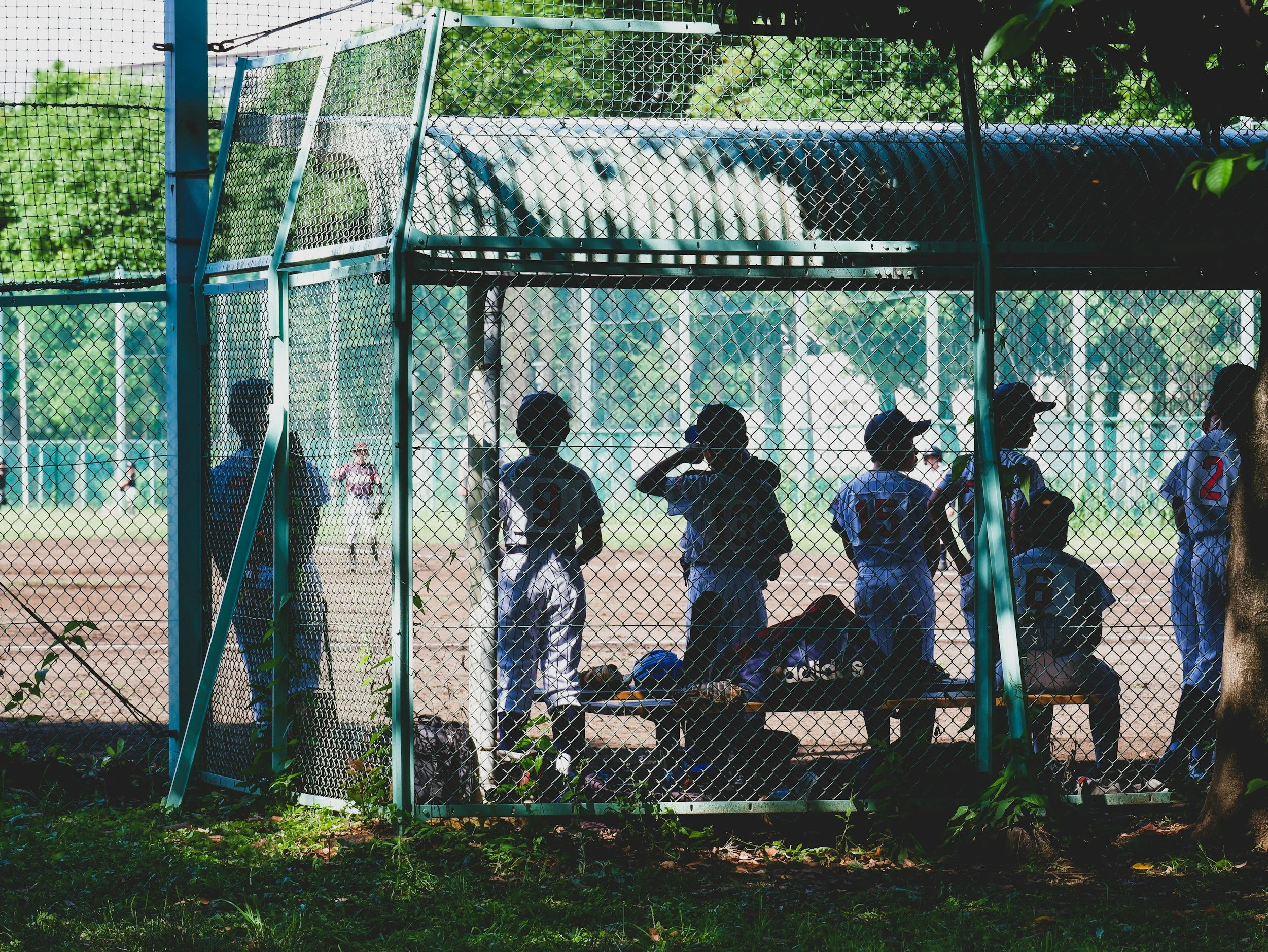 A group of boys in baseball uniforms standing and sitting inside a fenced baseball dugout, with a baseball field and other players visible in the background.