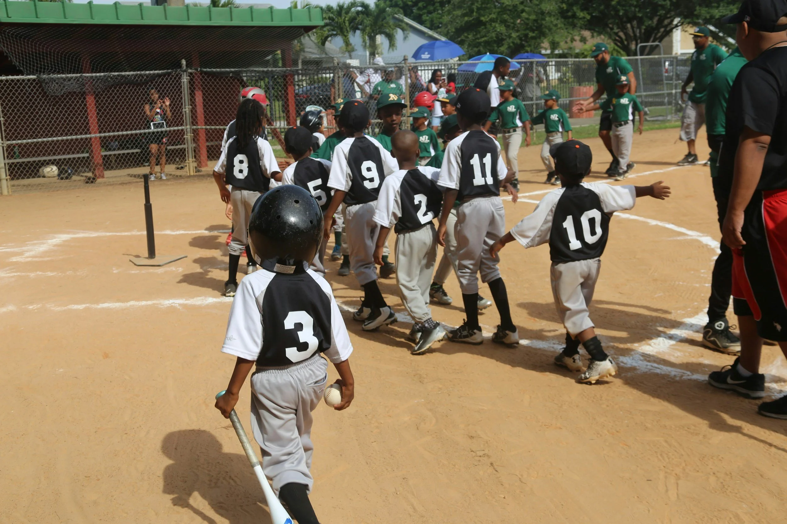 Young baseball players in black and white uniforms shaking hands after a game on the field with coaches and spectators in the background.