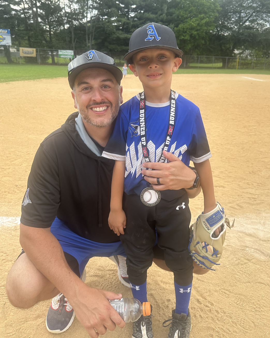 A man kneeling on a baseball field, smiling, with a young boy holding a baseball medal around his neck, both wearing baseball uniforms and hats, during daylight.