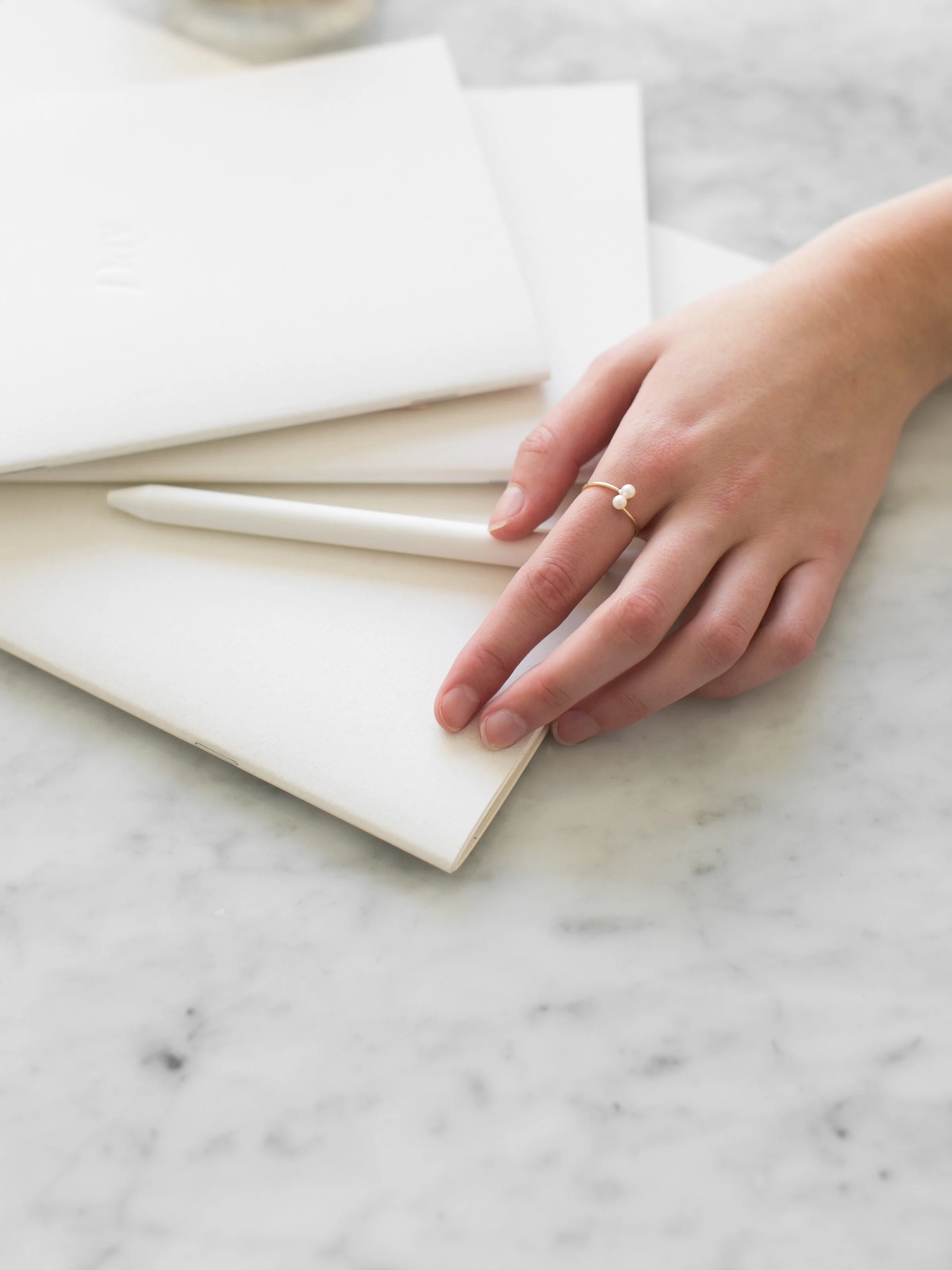 A hand with a pearl ring resting on a white notebook with a white pen, on a marble surface.