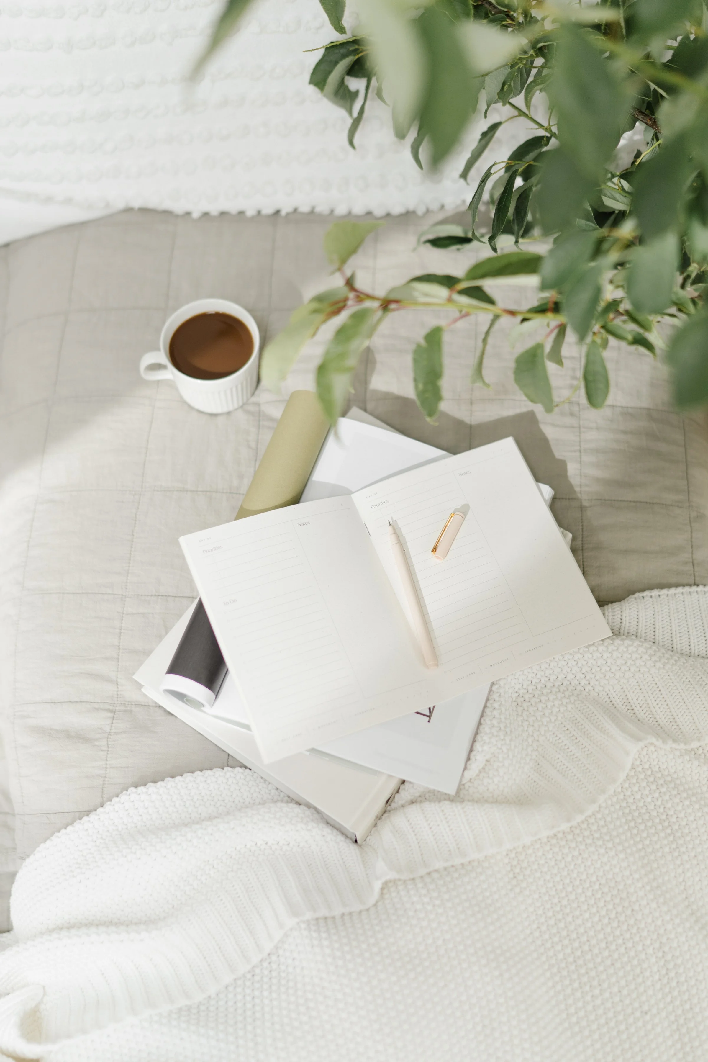 A cozy scene with a white knitted blanket, an open planner with lined pages, a white pen with a gold cap, a rolled-up magazine, a white mug filled with coffee, a green plant, and a white textured surface.