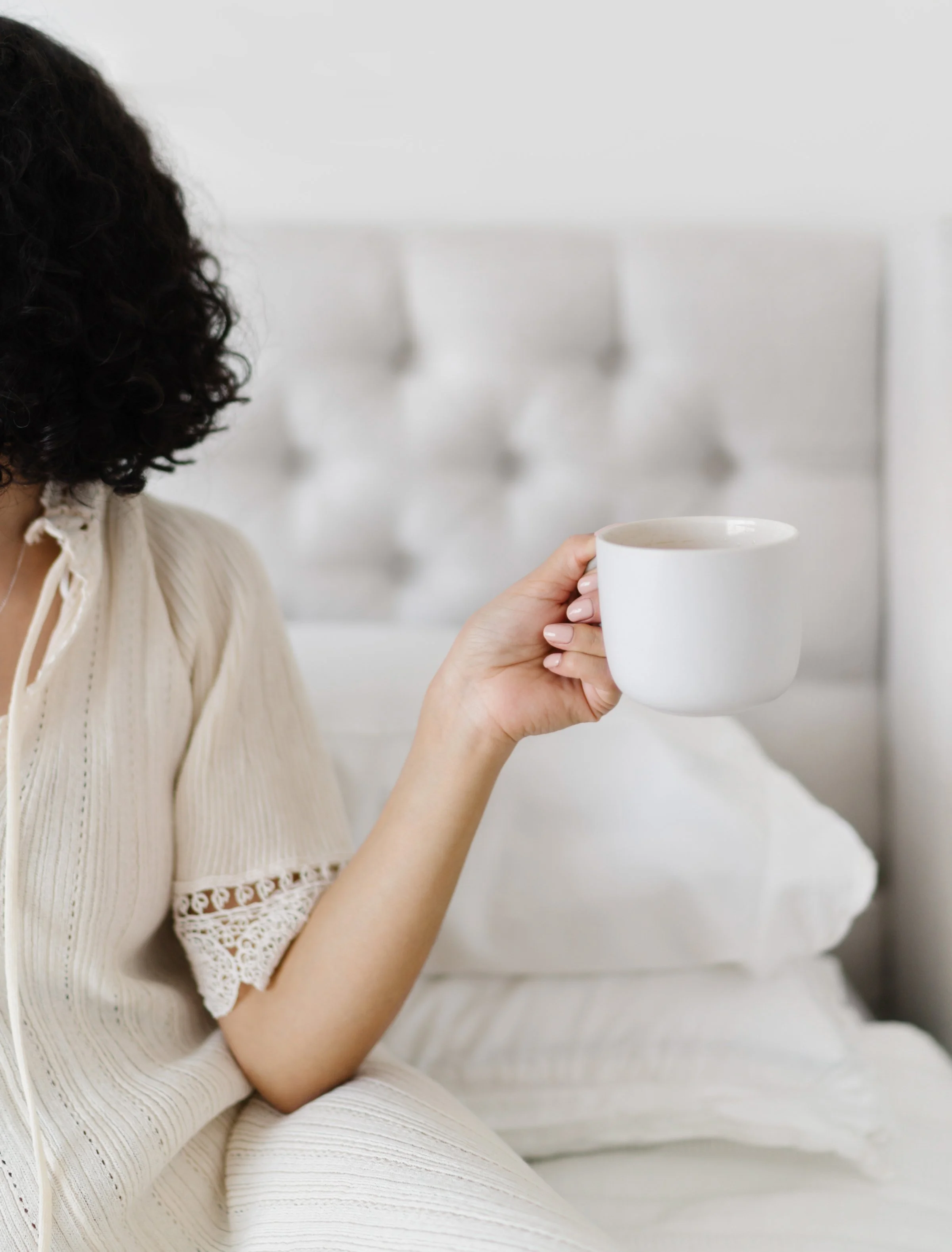 A woman dressed in a cream-colored, textured dress with lace accents on the sleeves, holding a white mug with her right hand. The background is blurred, showing a white tufted headboard or furniture.