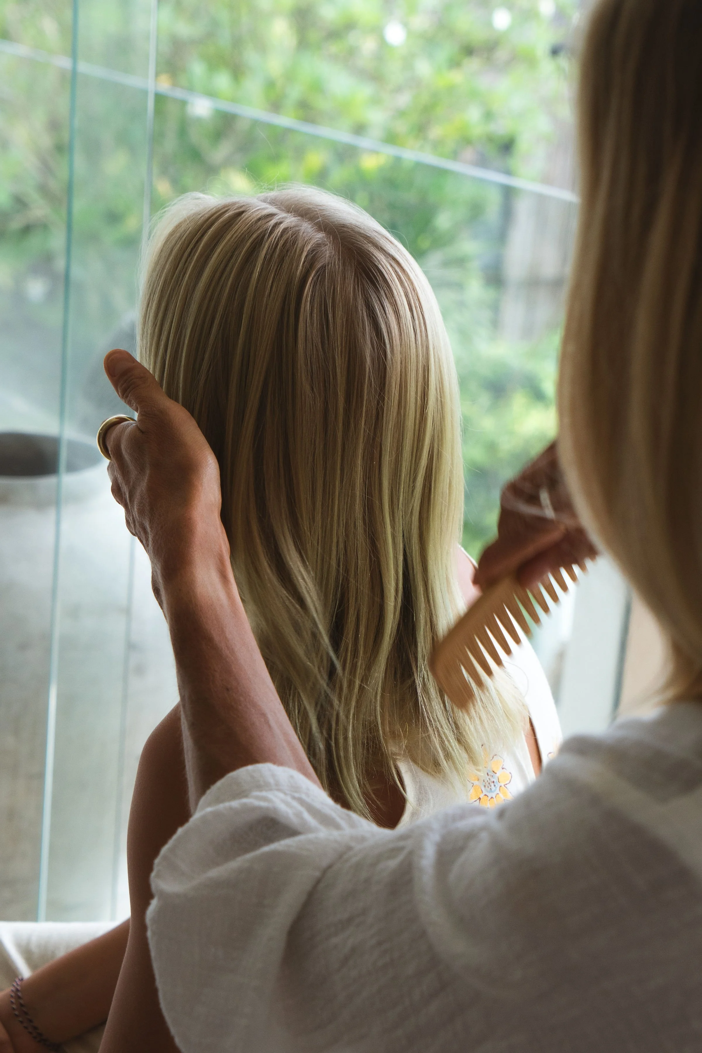 A person with light blonde hair and a white shirt combing a young girl's long blonde hair while she looks out a window with a green outdoor scene.