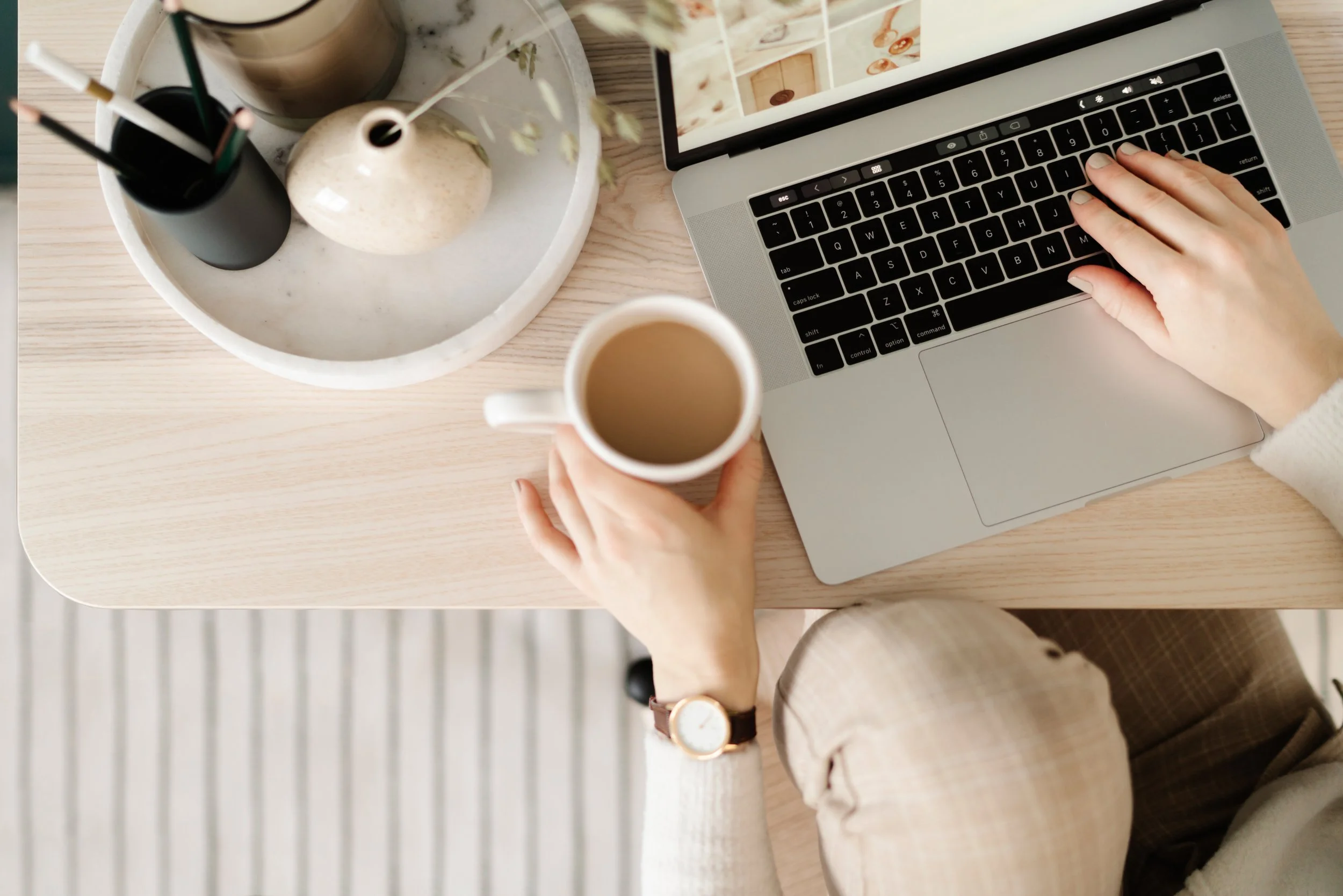 A person working on a laptop at a wooden desk, holding a cup of coffee in their right hand, with a watch on their wrist. The desk also has a white tray with some decorative items and a smartphone displaying images.