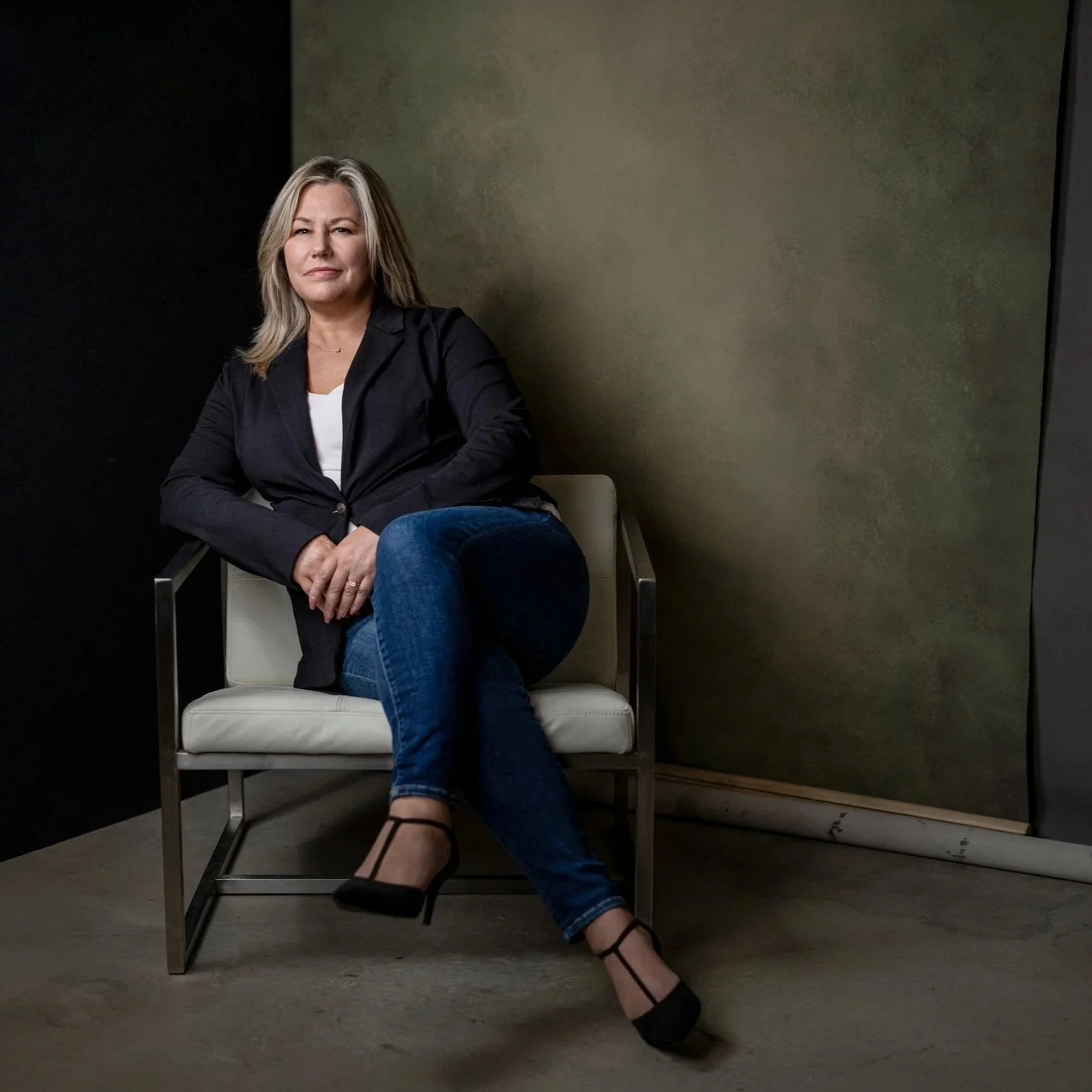 A woman with blonde hair sits on a modern chair in front of a two-tone wall, wearing a black blazer, white top, blue jeans, and black heels.