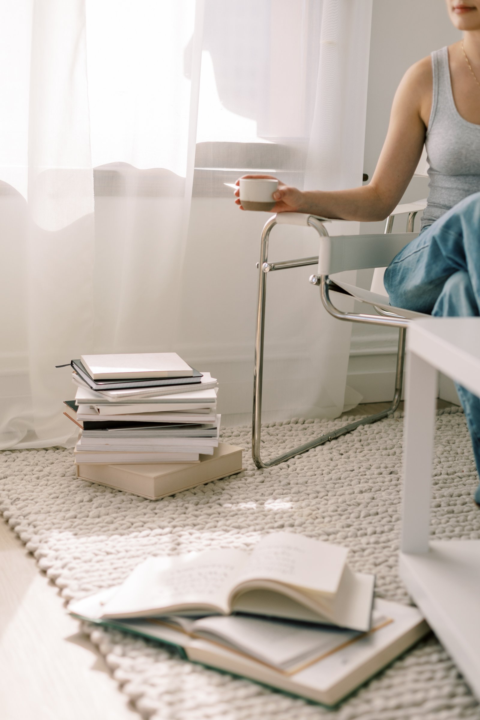 A woman sitting on a modern chair in a bright room, holding a cup. In the foreground, there are stacked notebooks and open books on a textured rug.