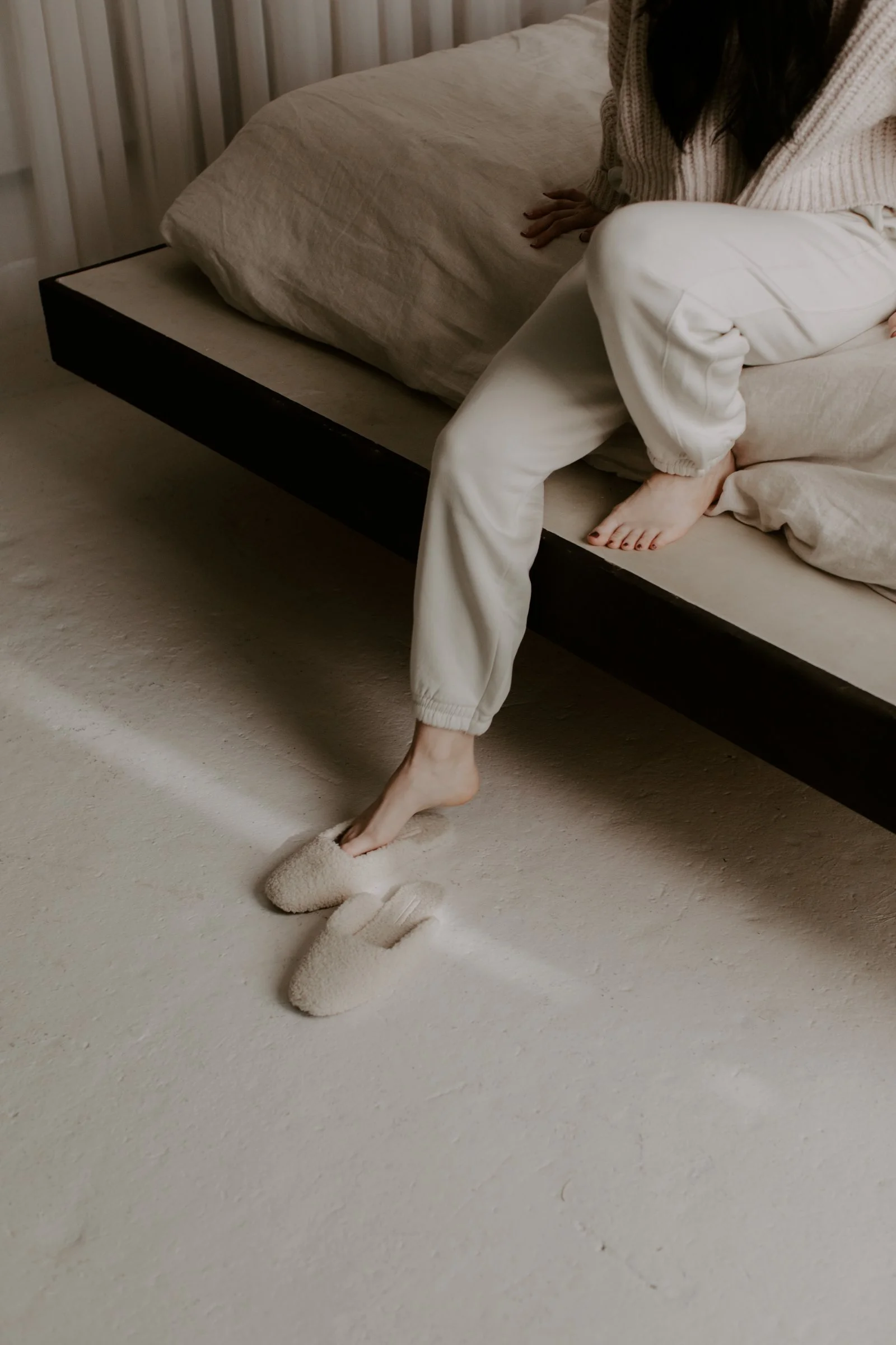 A woman sitting on the edge of a bed with beige linens, wearing cream-colored sweatpants and slippers, with one foot on the floor.