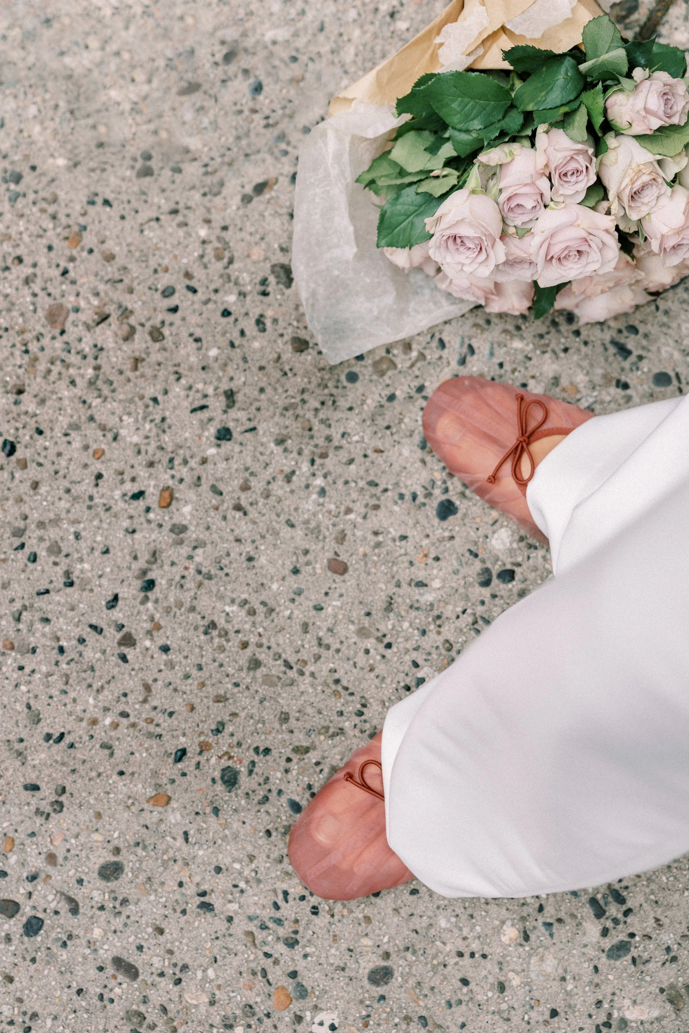 A person in beige shoes and white pants kneeling on a pointed knee on a gravel ground, next to a bouquet of pink roses wrapped in brown paper.