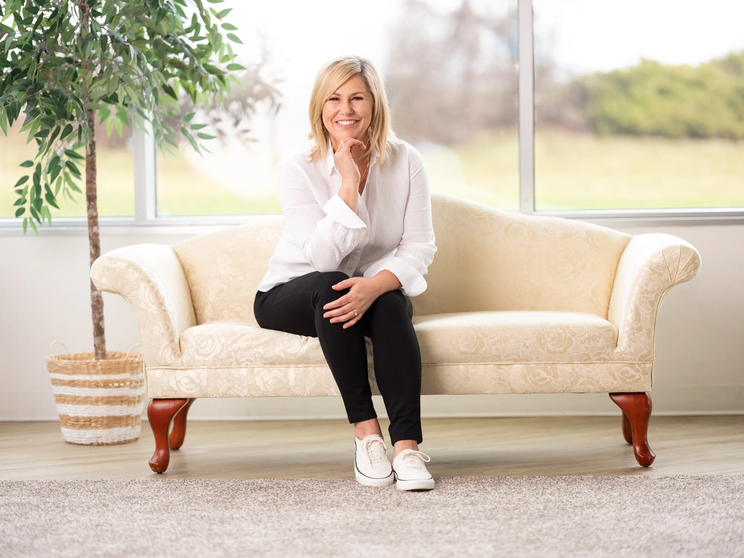 A woman with blonde hair wearing a white shirt and black pants sitting on a cream-colored vintage sofa in a bright room with large windows, green plants, and a view of the outdoors.