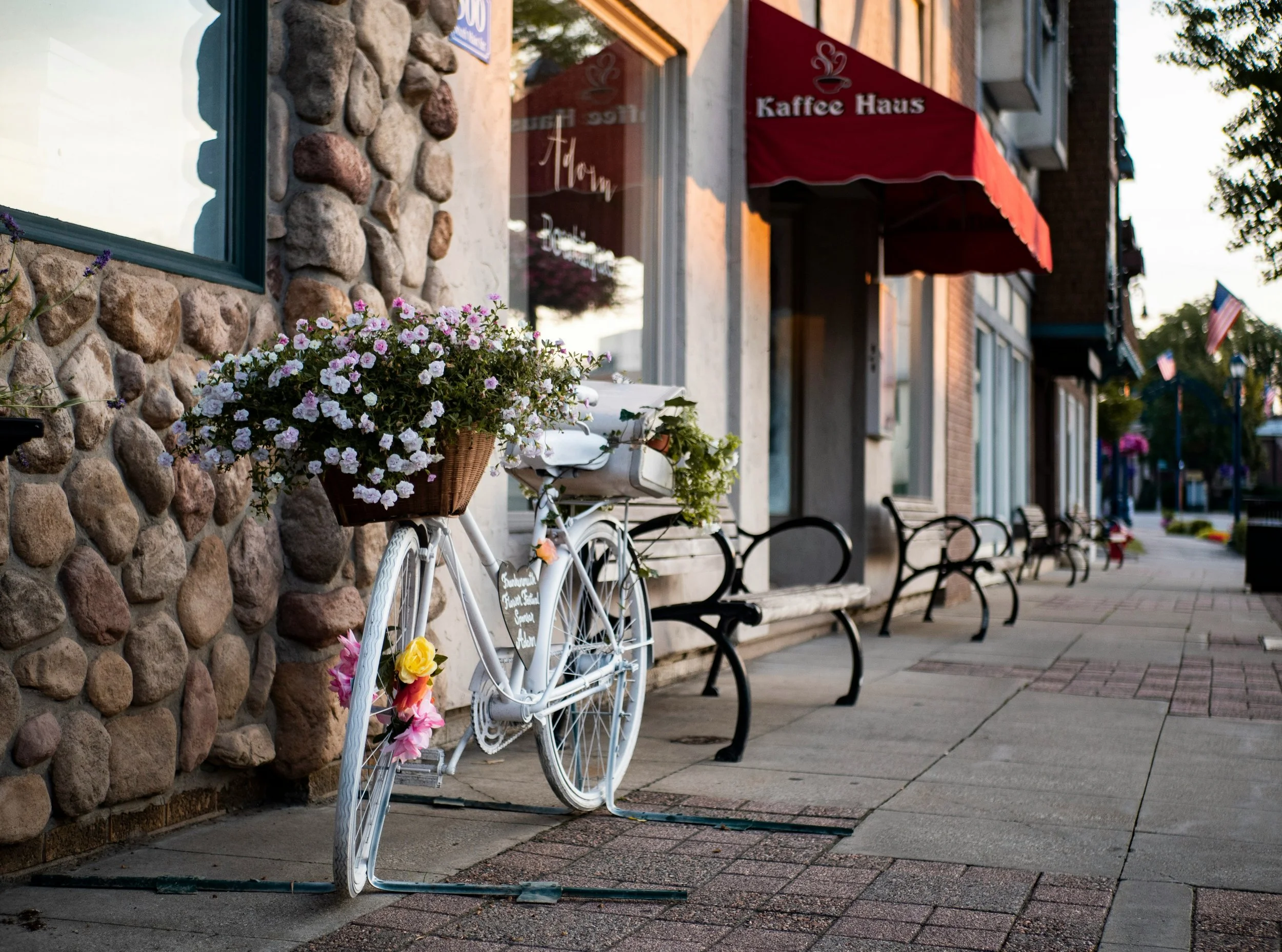 A white vintage bicycle with a basket of pink and white flowers parked on a sidewalk outside a cafe in Frankenmuth, Michigan with a stone and brick facade, red awning, and benches.