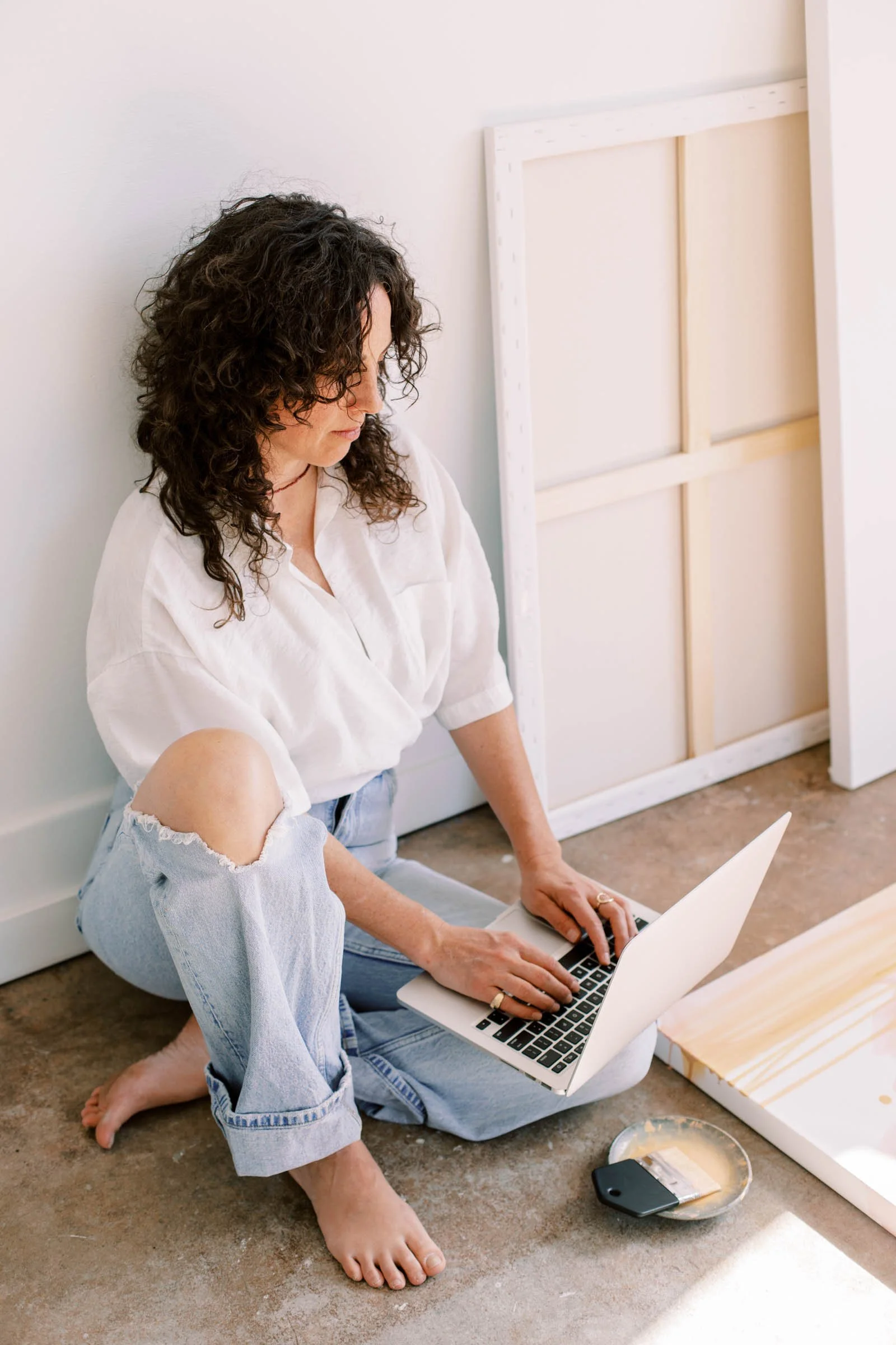 A woman with dark curly hair and a white shirt is sitting on the floor, working on a laptop. She is barefoot and wearing ripped jeans. There is an art canvas propped against the wall behind her, a paint palette and a brush on the floor, and a paint roller next to her.