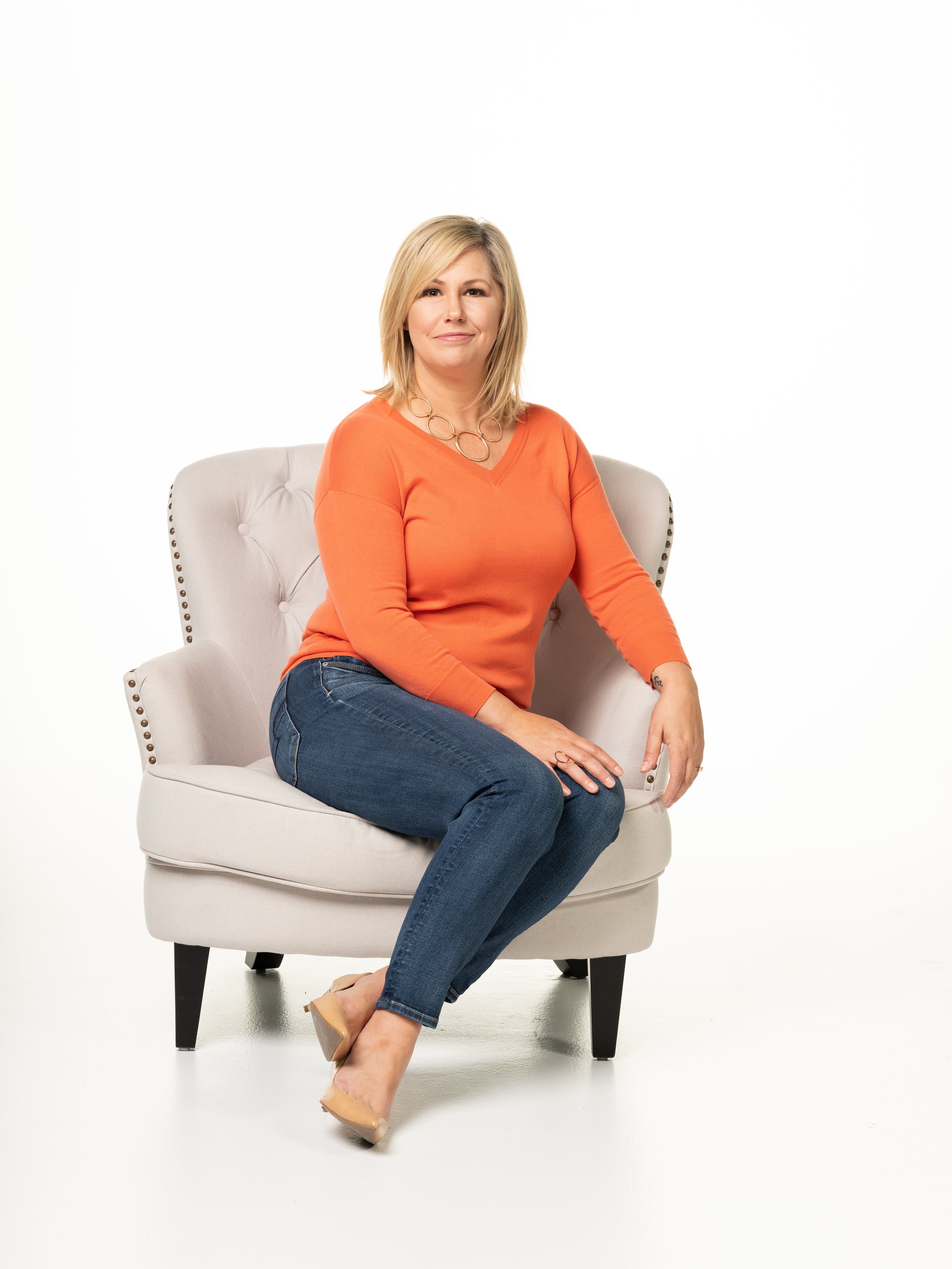A woman with blonde hair wearing an orange long-sleeve top, blue jeans, and nude heels, sitting on a beige armchair with black legs against a white background.