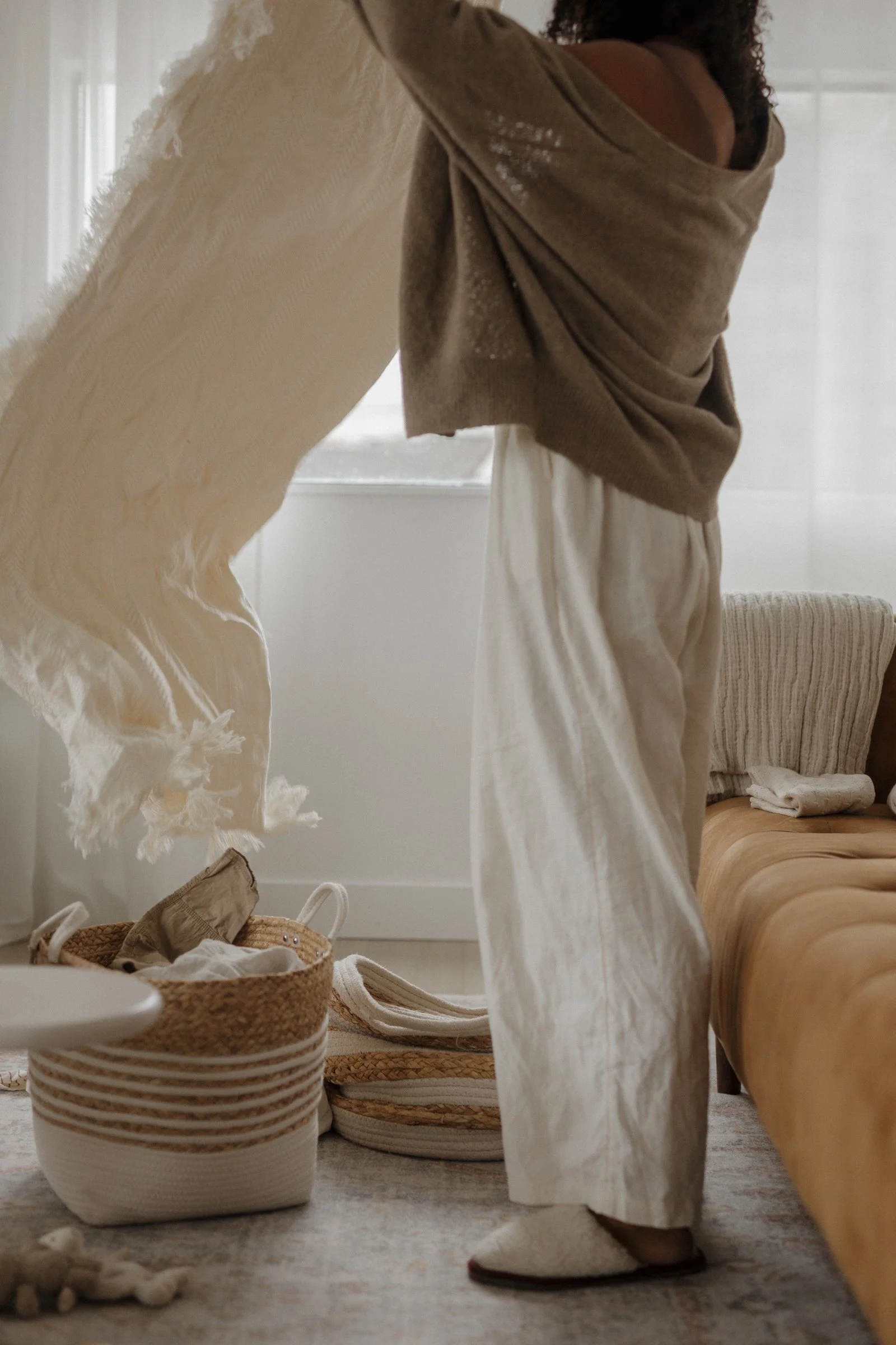 Person wearing loose beige pants and slippers, standing in a softly lit room with a beige sofa, a woven basket, and shoes on the floor.