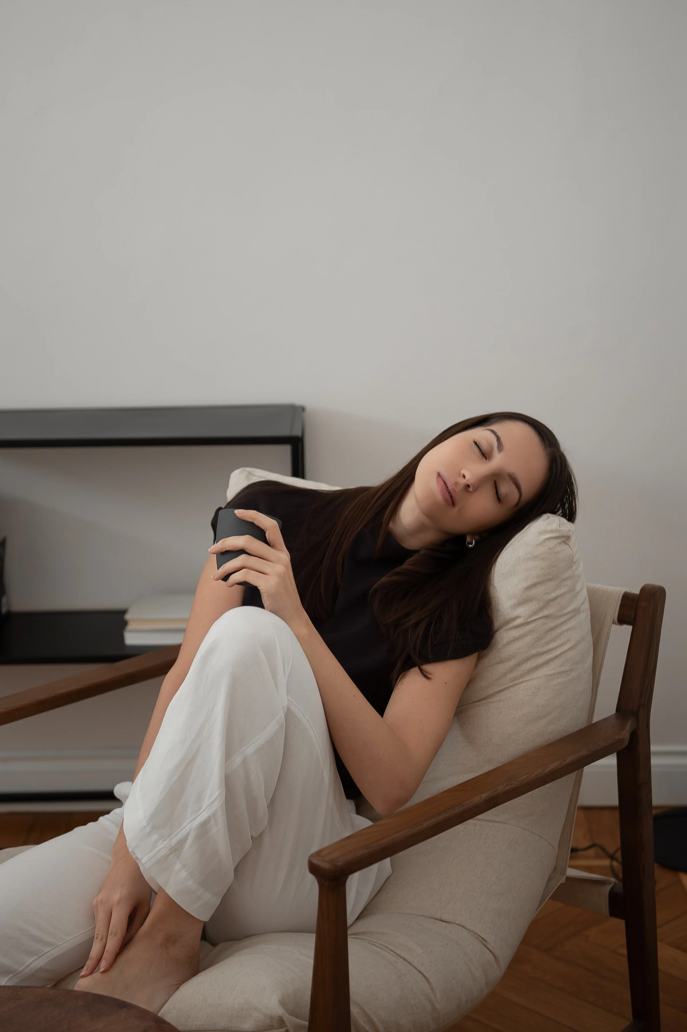 A young woman with long brown hair peacefully sleeping on a beige chair, holding a black mug, in a room with white walls and minimal decor.
