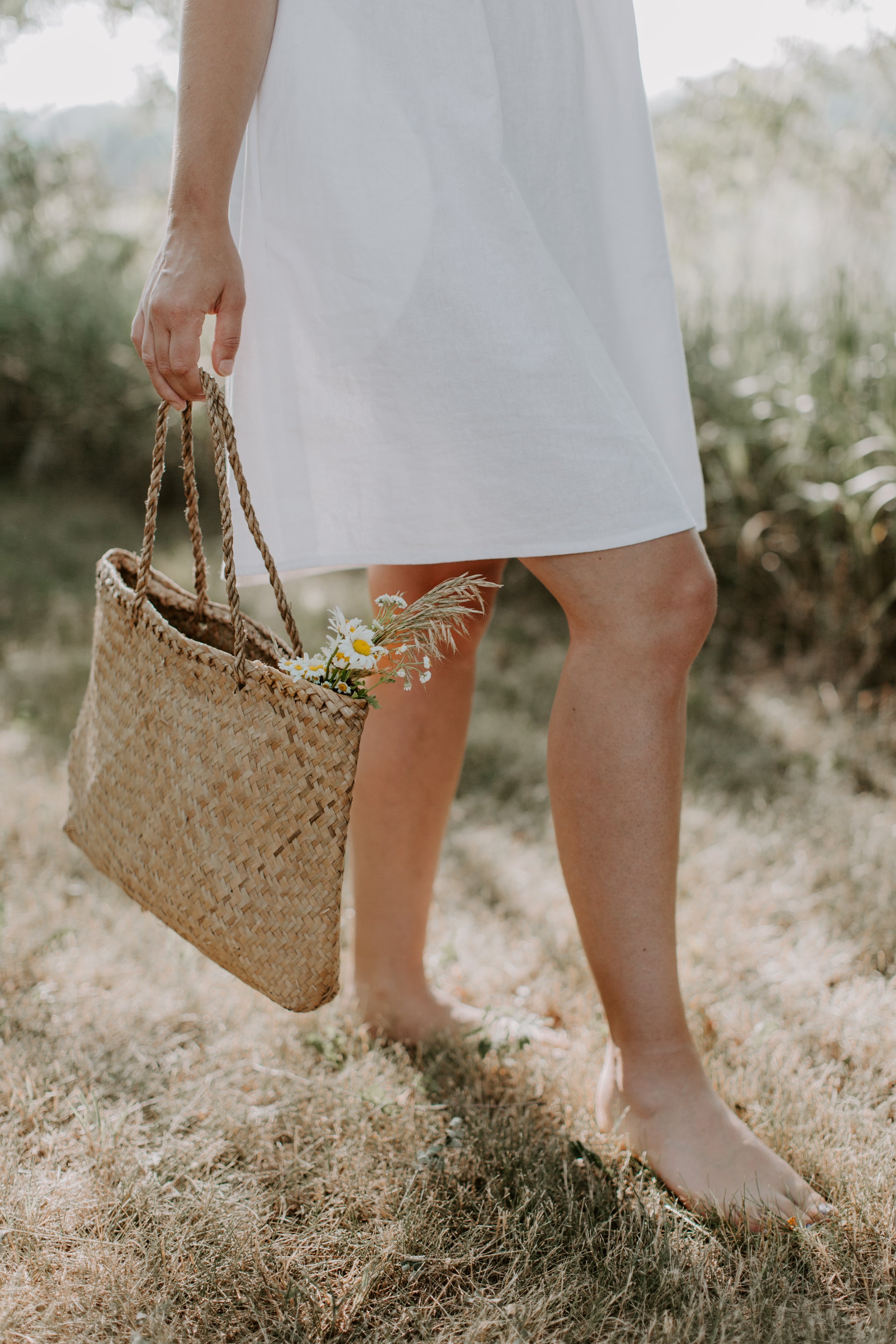 Person in a white dress stands barefoot on dry grass, holding a woven bag with flowers in it.