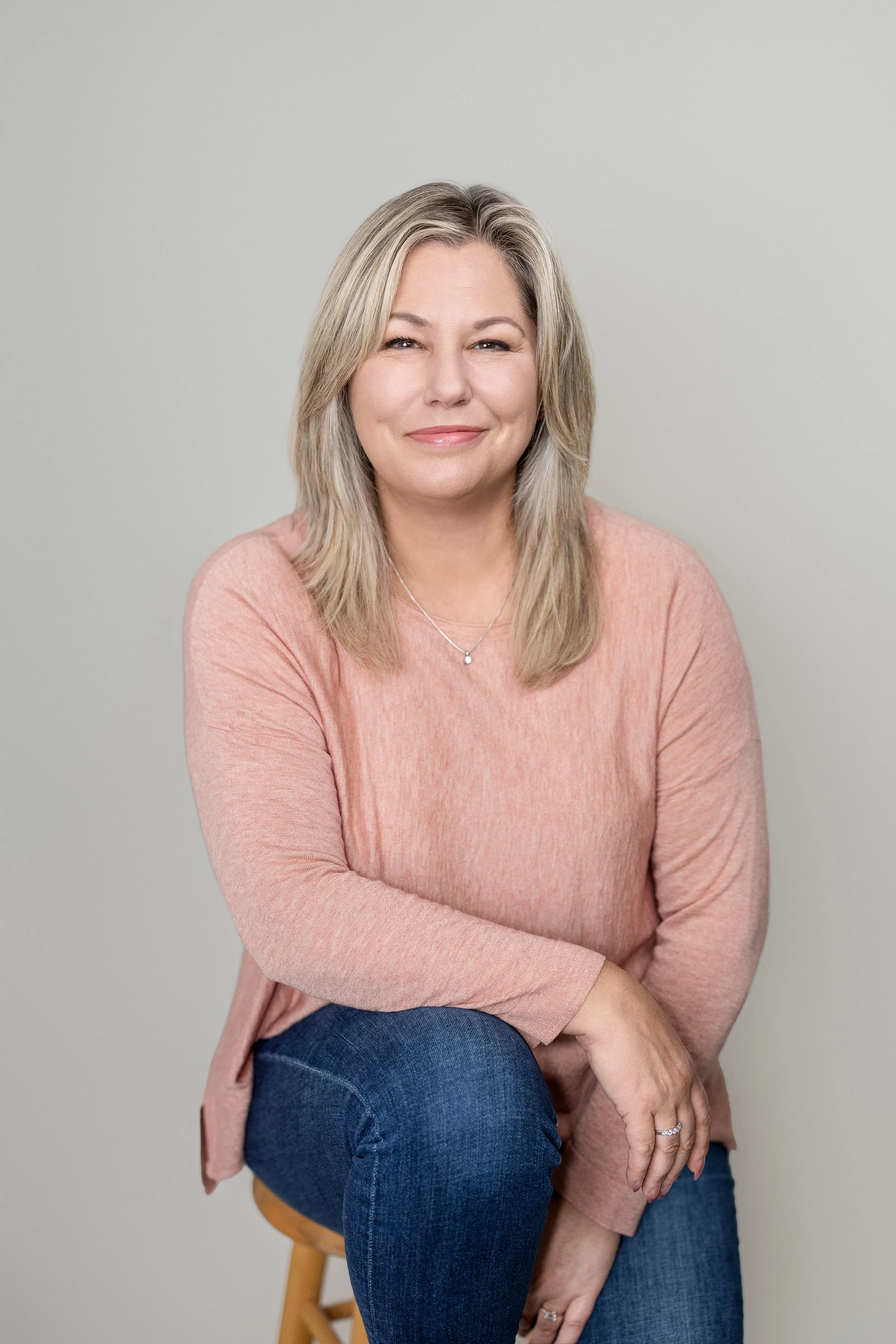 A woman with shoulder-length blonde hair, wearing a peach-colored long-sleeve top and blue jeans, sitting on a stool against a plain light background.