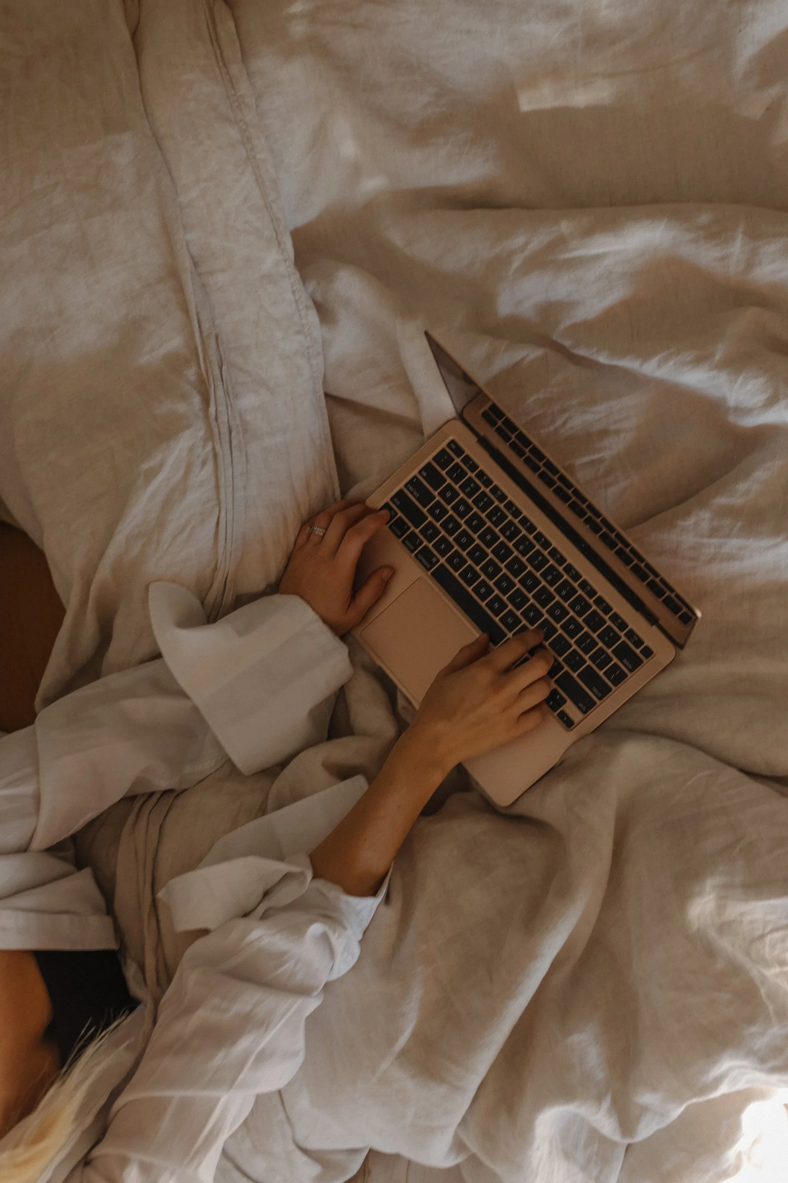 Person using a laptop on a bed with beige sheets, wearing a white long-sleeve shirt and beige shorts.