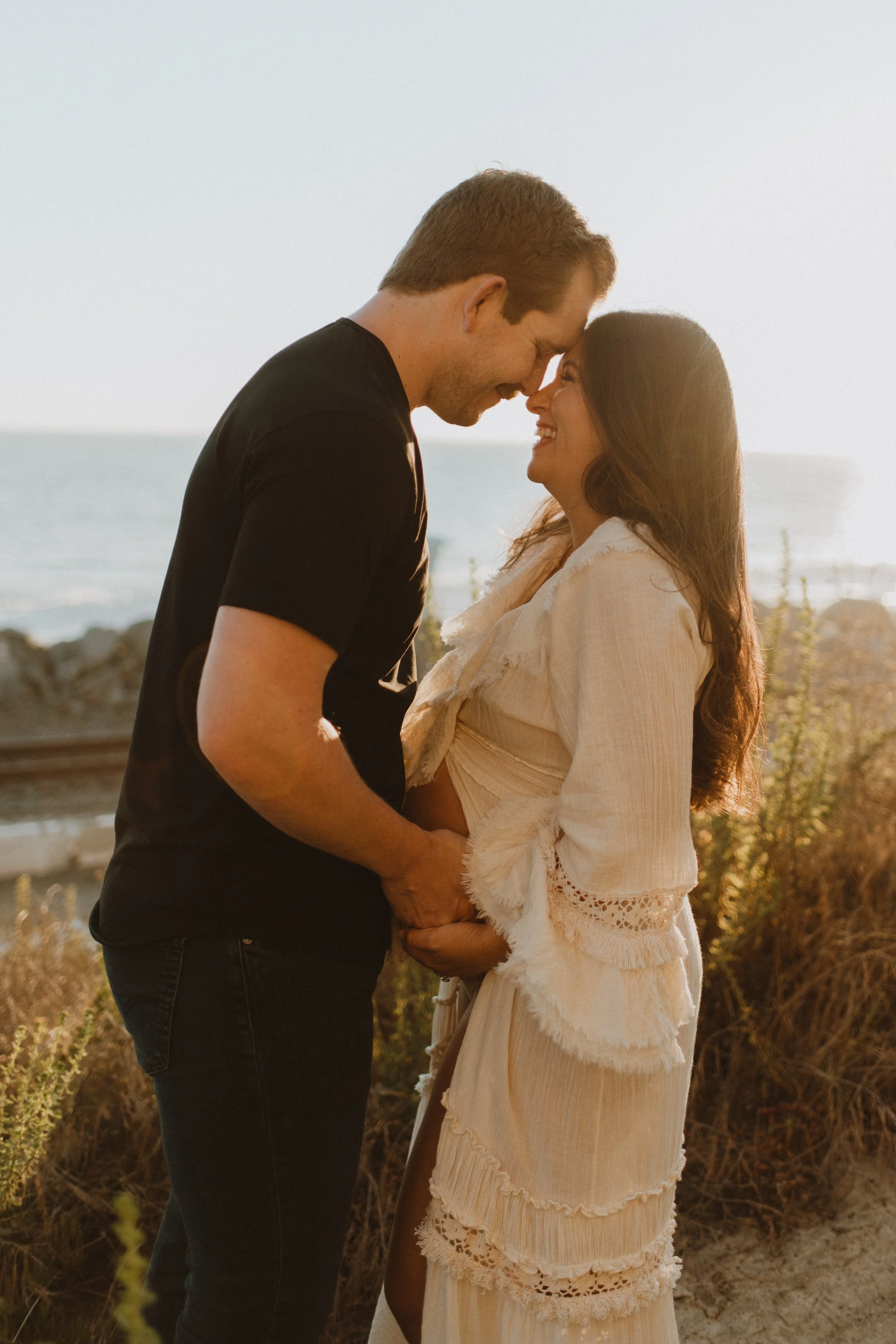 A couple standing close together on a beach at sunset, smiling and touching foreheads, with the ocean in the background.