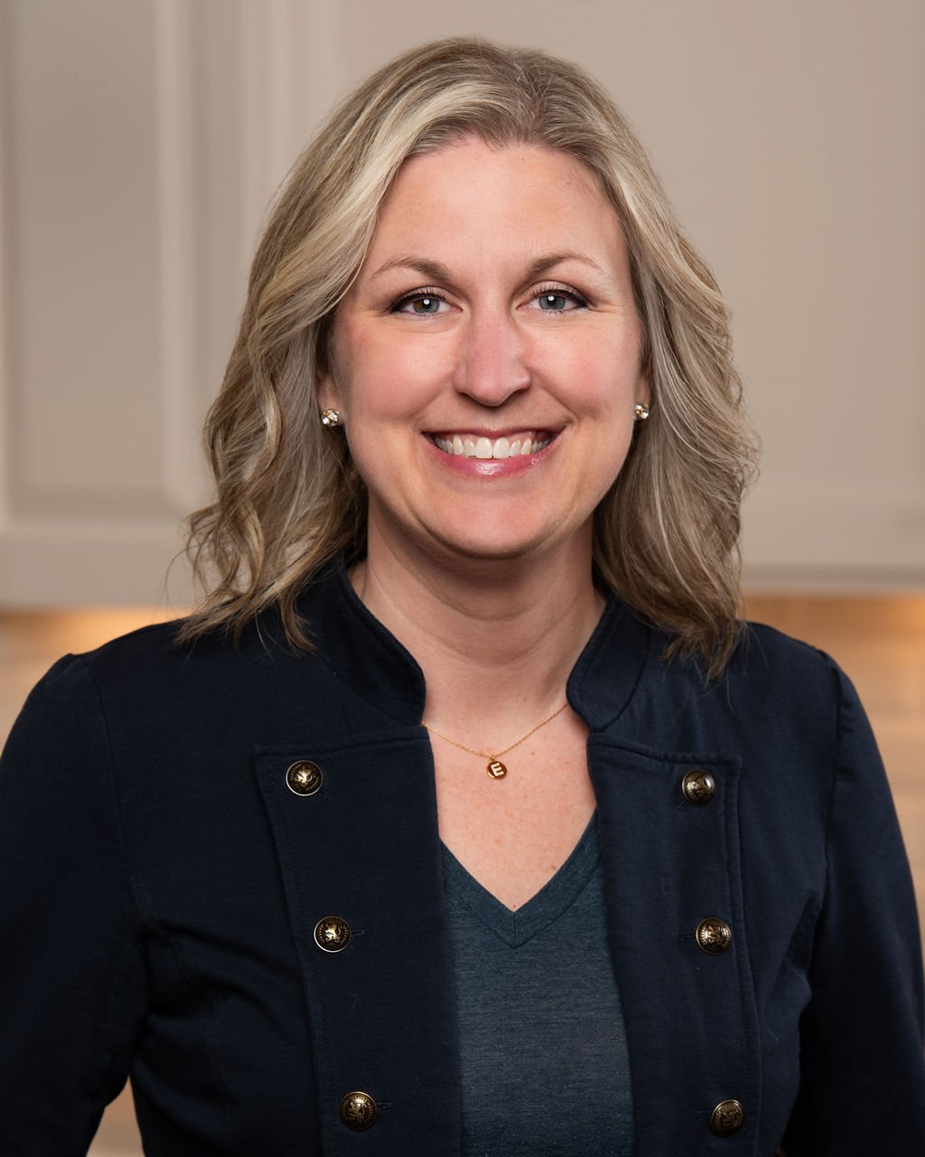 A smiling woman with blonde wavy hair, wearing a dark navy jacket with gold buttons, a gray top, gold earrings, and a gold necklace with a small pendant.