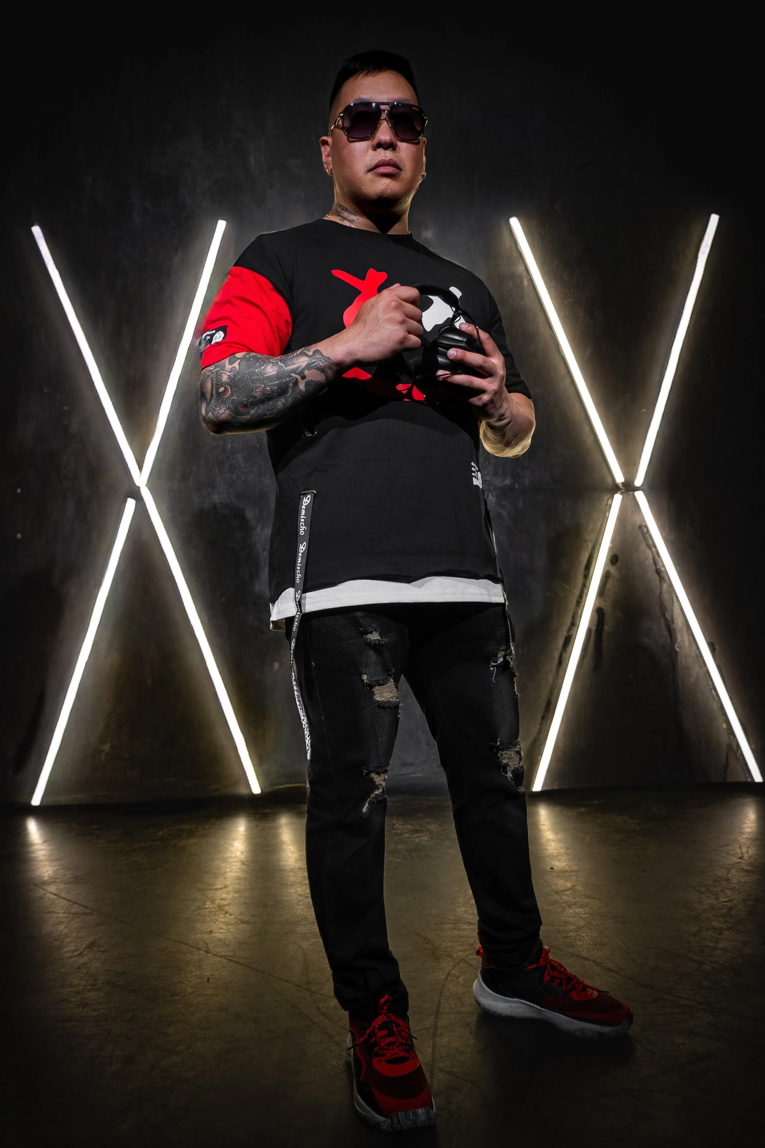 A man wearing sunglasses, a black and red t-shirt, ripped jeans, and red sneakers stands in front of a black wall with neon light tubes forming an 'X' shape behind him, holding headphones.