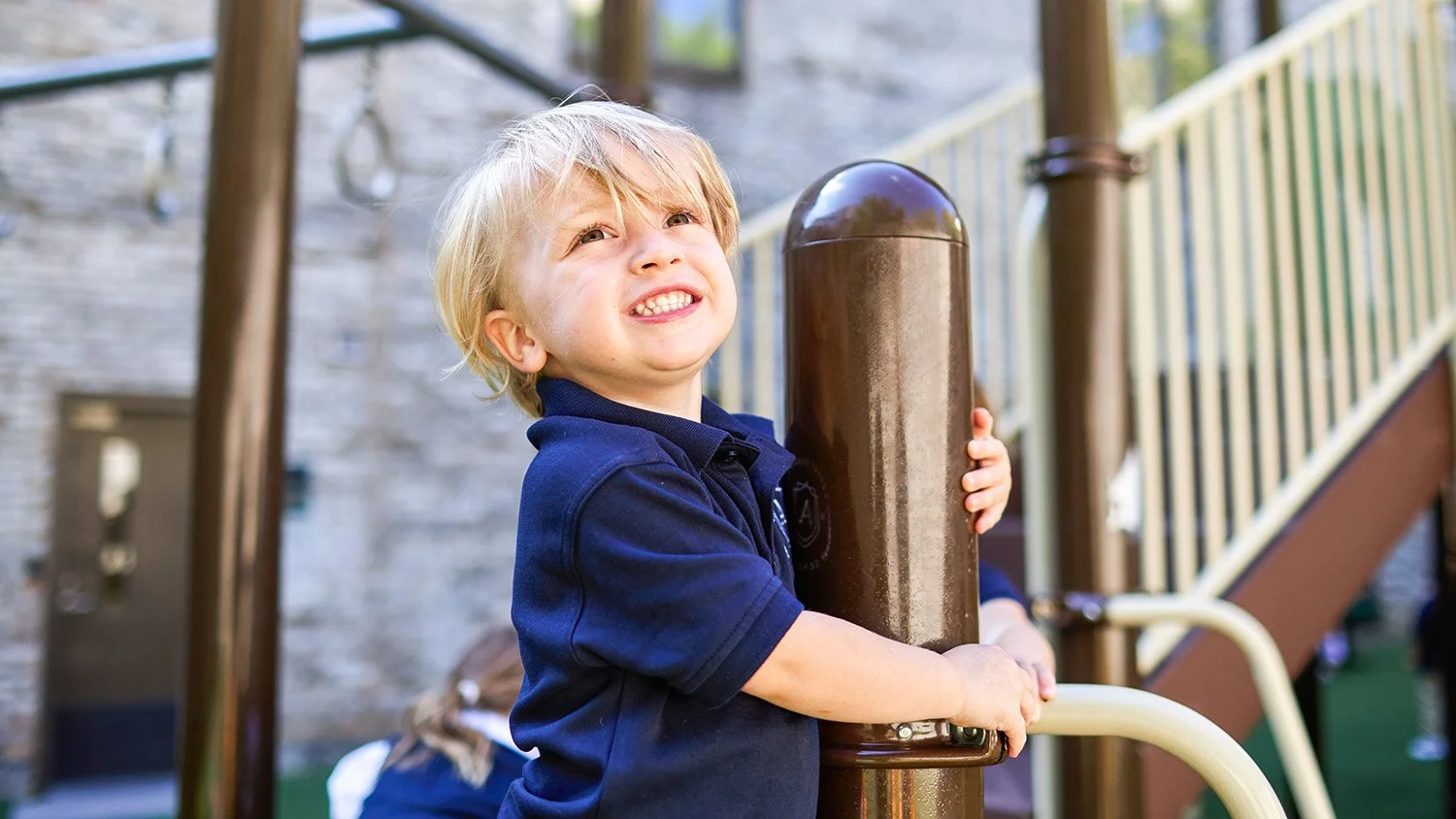 A young boy with blond hair, wearing a navy blue shirt, is smiling and holding onto a brown playground pole.