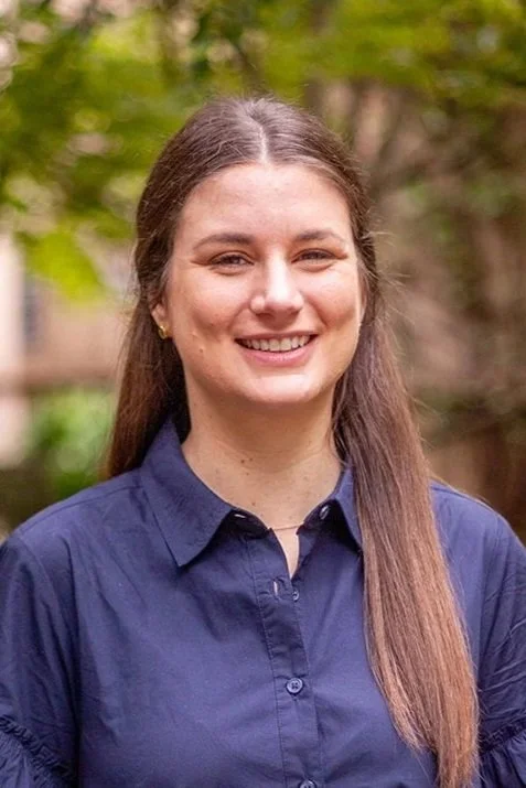 Portrait of a smiling woman with brown hair, wearing a navy blue shirt and a gold necklace, standing outdoors in front of green foliage.