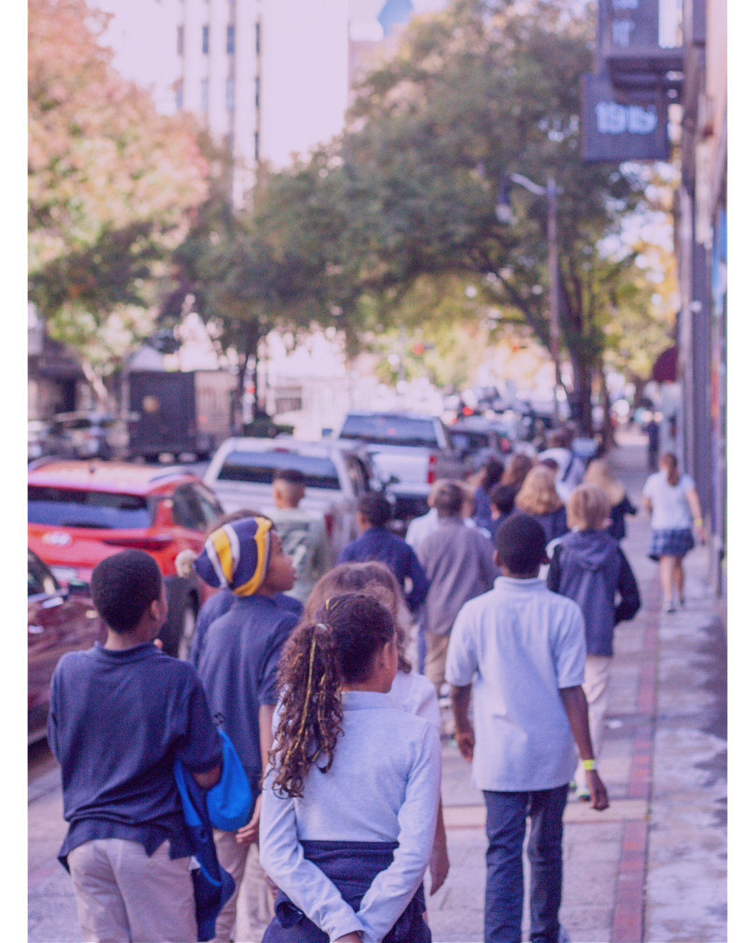 A group of children walking on a city sidewalk during daytime, with parked cars and trees lining the street.