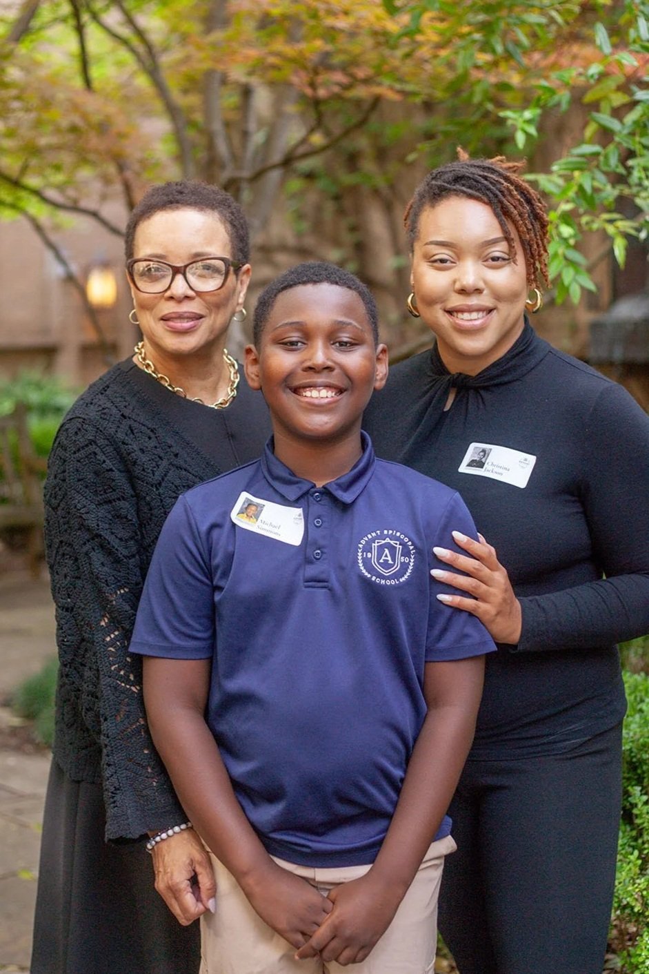 A woman, a young boy, and another woman pose together in an outdoor setting with greenery and trees in the background. The woman on the left wears glasses, a black sweater, and gold jewelry. The woman on the right has short, styled hair, and wears a black top. The boy in the middle wears a blue school uniform polo shirt with a logo and a name tag. All three are smiling and standing close together for a picture.