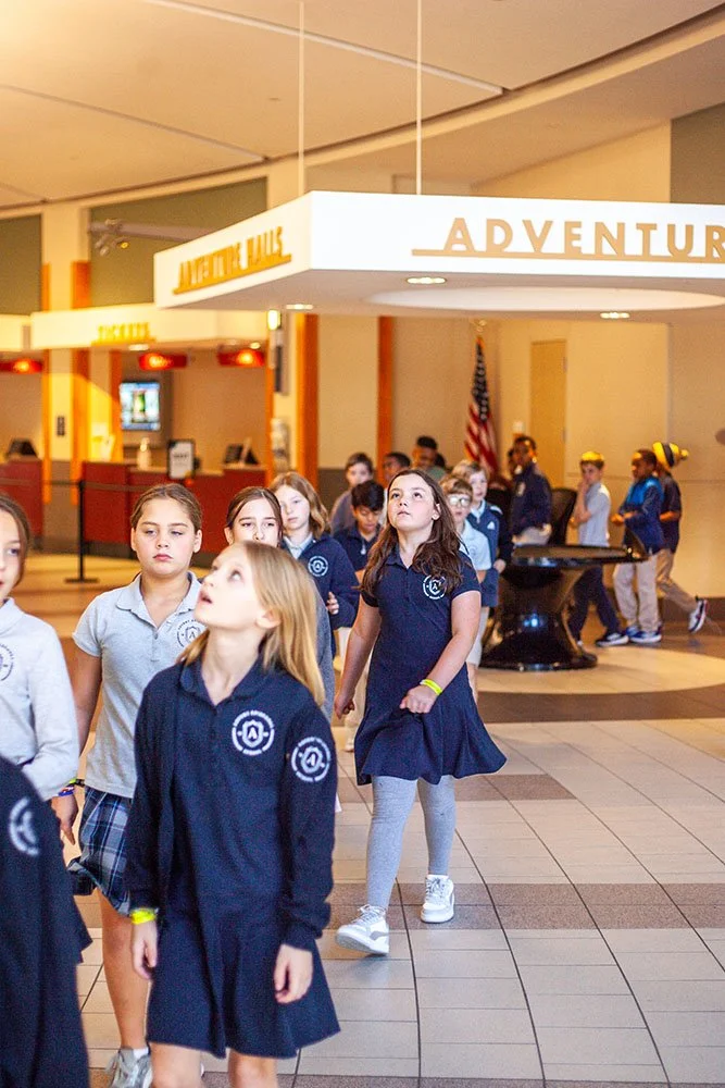 Group of children in school uniforms walking through an indoor recreational or entertainment center with a sign that reads "Adventure Valley" in the background.