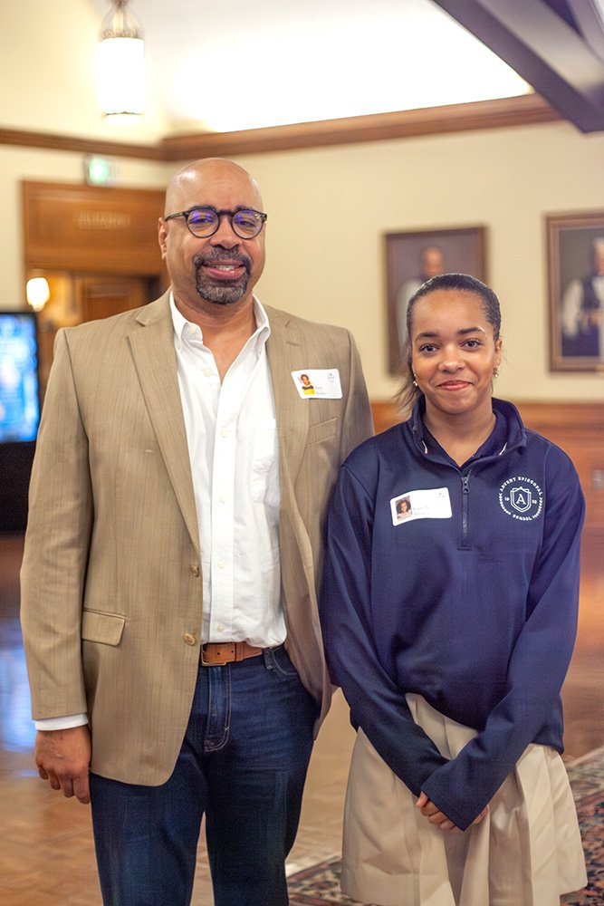 A man in a beige blazer, white shirt, and glasses standing next to a girl in a navy blue jacket and beige skirt in an indoor setting with framed photos on the wall.