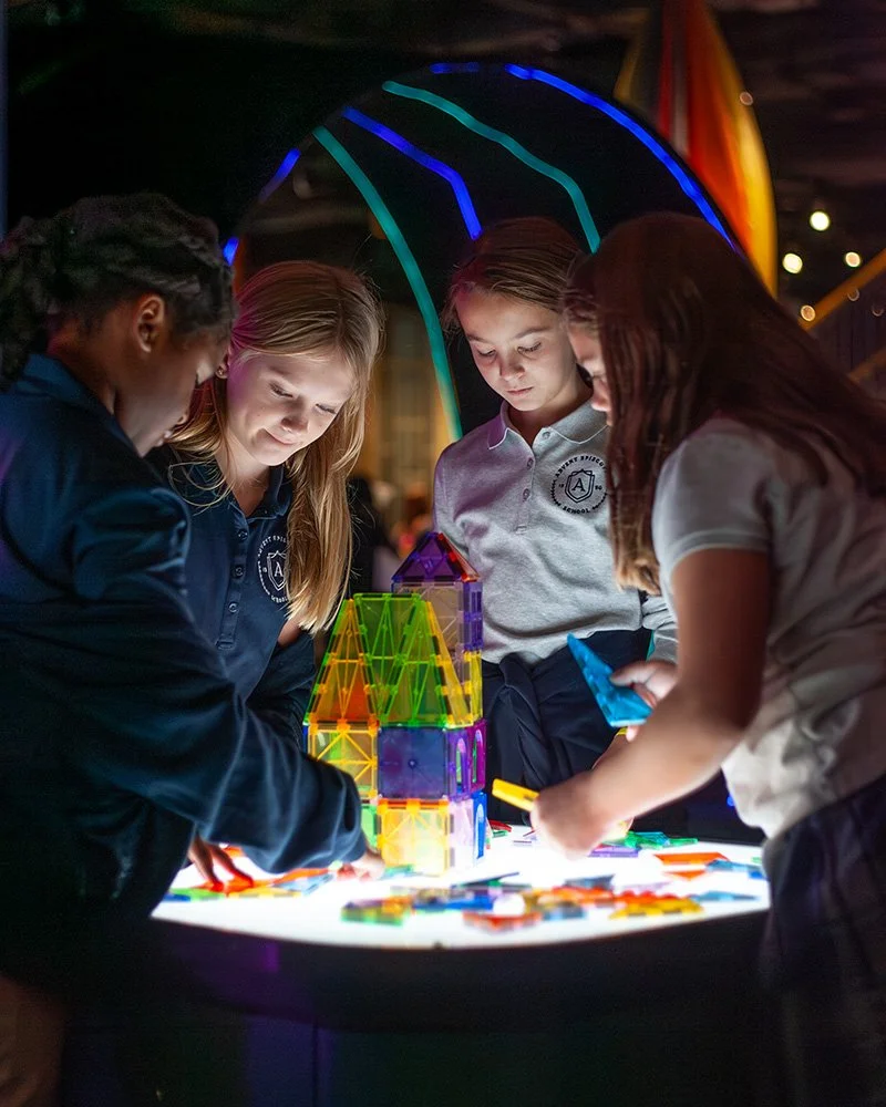 Four children gathered around a glowing table playing with colorful magnetic building tiles at a science exhibit at McWane Science Center.