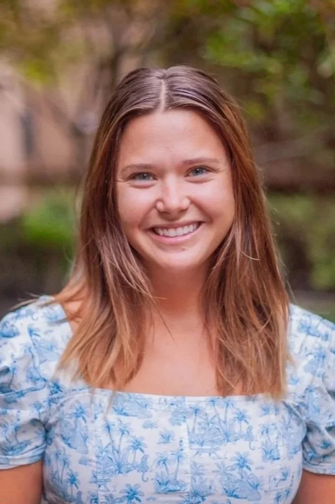 A young woman with shoulder-length blonde hair, blue eyes, and a bright smile, wearing a floral-patterned top, standing outdoors in a green garden with lush foliage in the background.