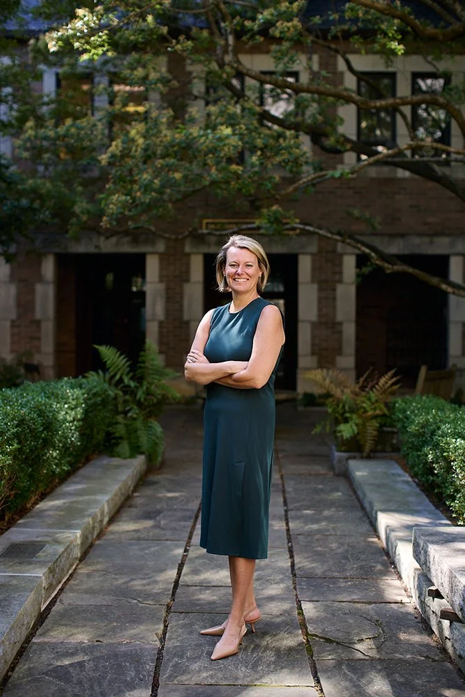A woman standing on a stone pathway outdoors, crossing her arms and smiling, with greenery and a brick building in the background.