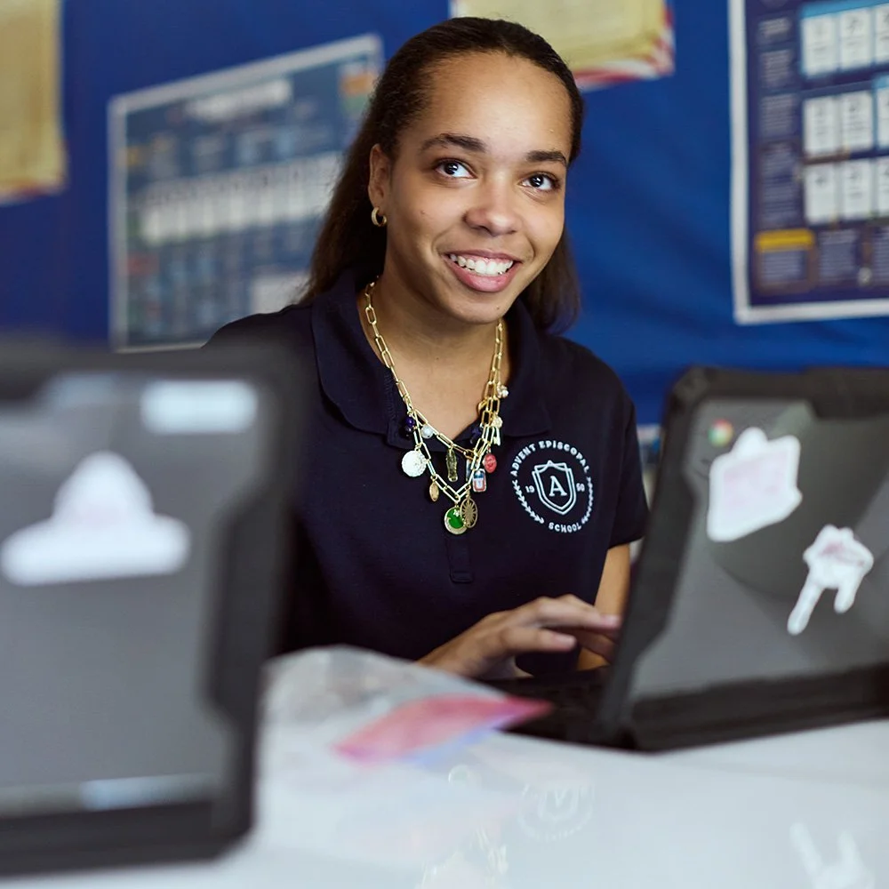 A young girl sitting at a desk with two laptops in front of her, smiling at the camera.