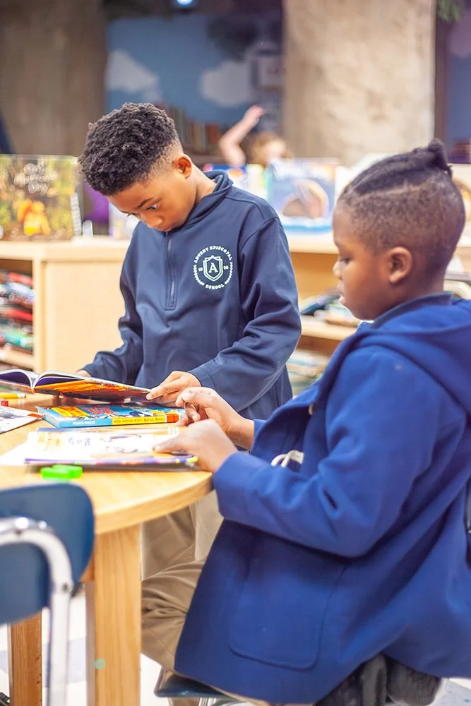 Two young boys with dark skin and curly hair, wearing blue school jackets, are engaging with books and stationery on a wooden table in a classroom.