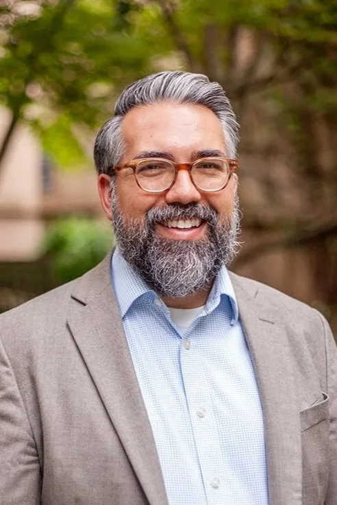 A smiling man with glasses, a beard, and gray hair, wearing a light blue blazer and a checked shirt, standing outdoors with green foliage in the background.