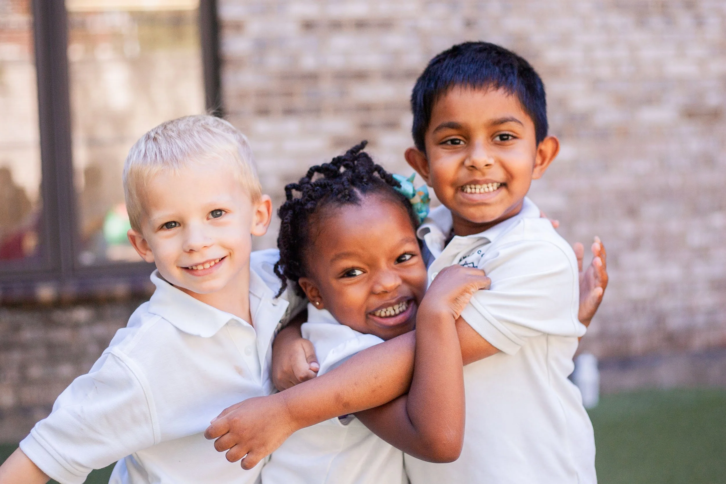 Three children hugging and smiling outdoors in front of a brick wall.