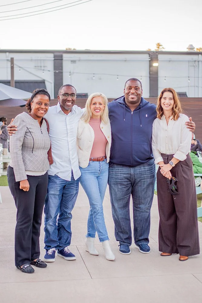 Group of five diverse adults smiling and posing together outdoors during daytime.